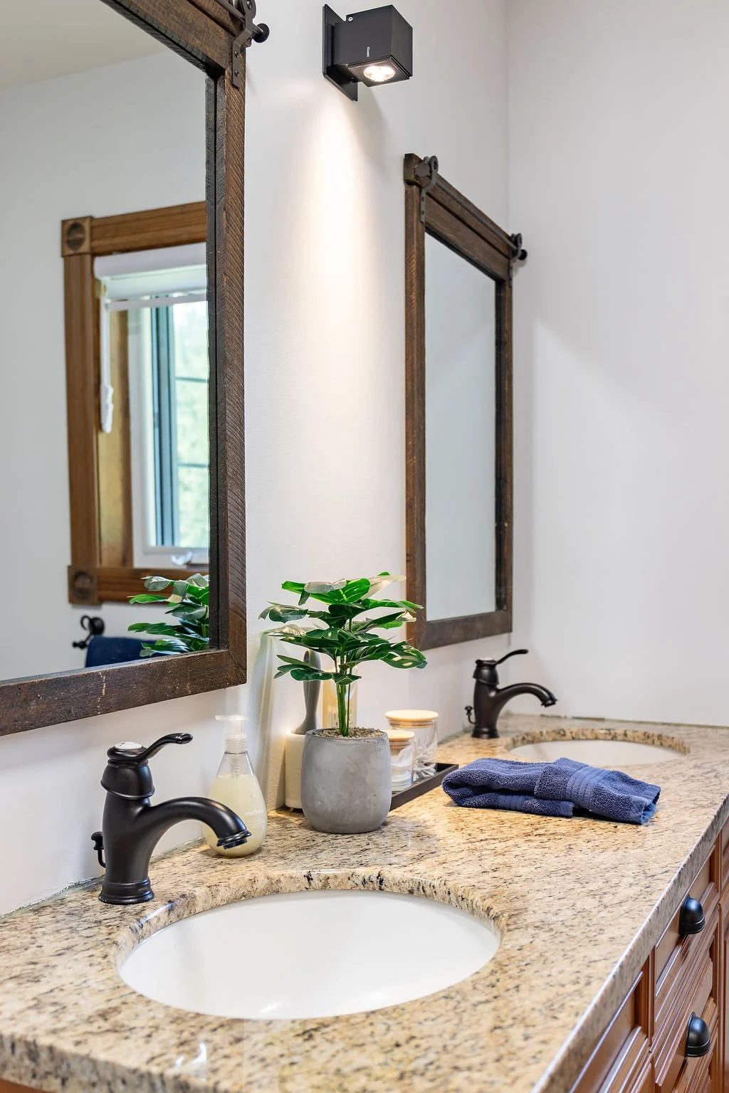 Bathroom vanity with granite countertop, two black faucets, two mirrors with wooden frames, a potted plant, soap dispenser, and folded blue towels.