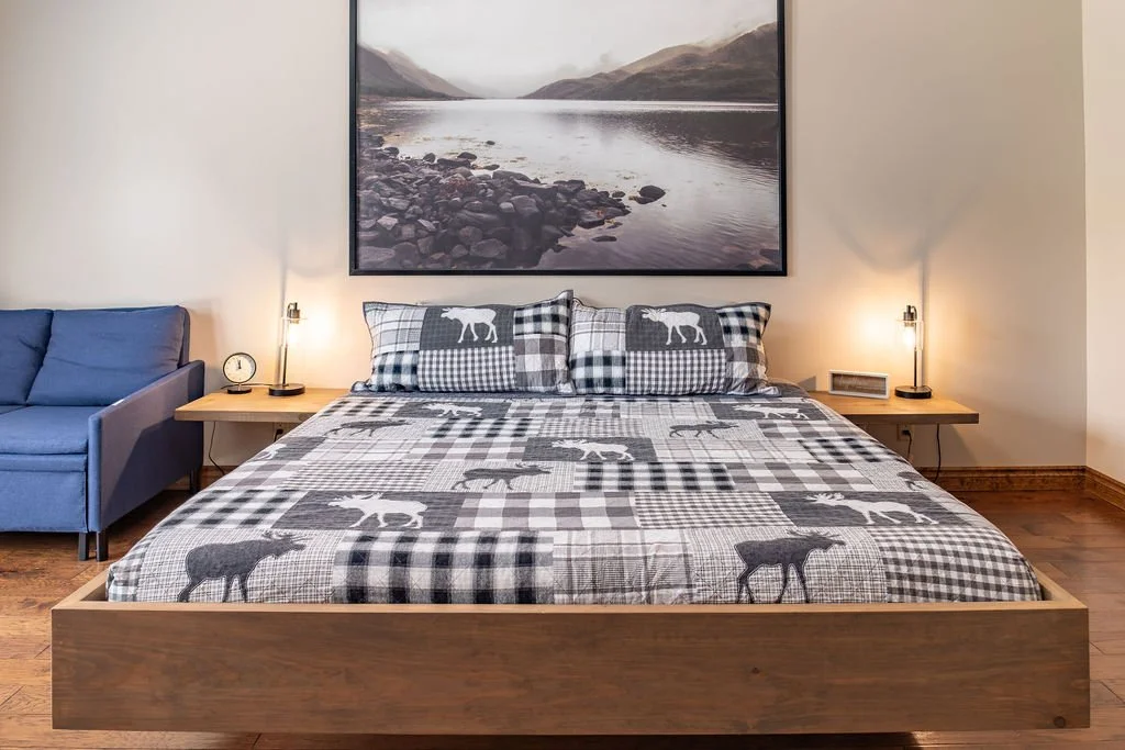 A bedroom with a wooden bed frame, a bed with moose-themed checkered bedding, two matching pillows, side tables with lamps and a clock, and a large landscape painting of a river and mountains hanging above the bed.