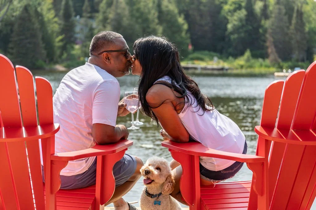 A couple sharing a kiss near a lake, seated in red chairs with a small dog in front of them, surrounded by trees.