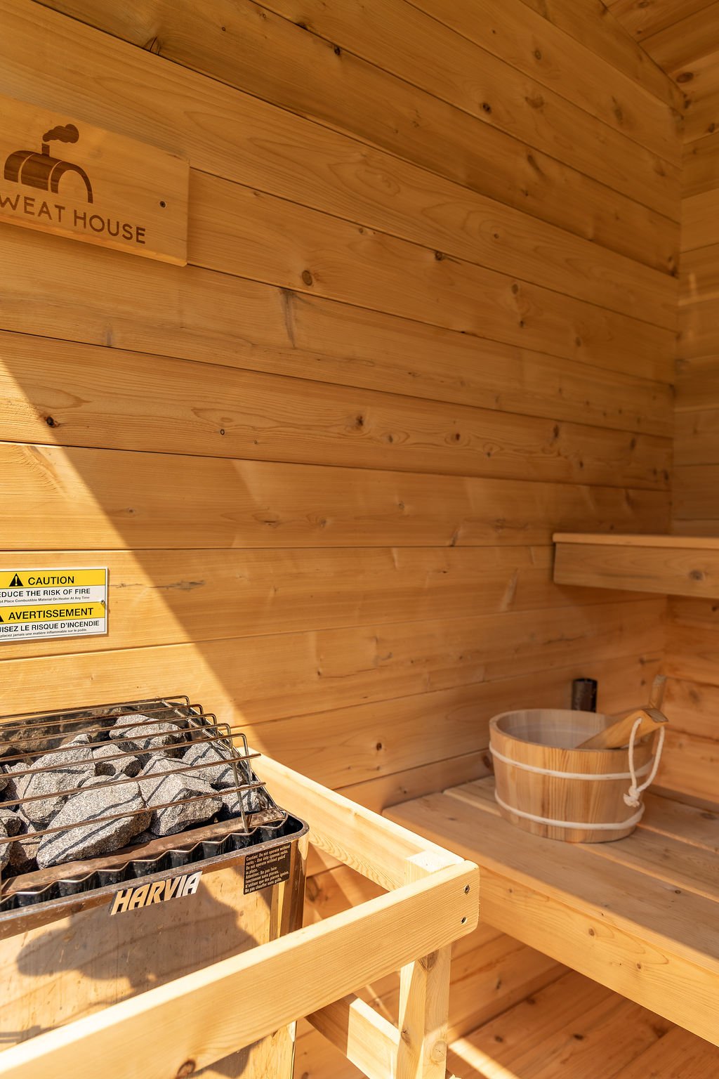 Wood interior of a sauna with a bucket and a ladle, a wooden sign with a chimney icon labeled 'Weather House', and a yellow caution sign.