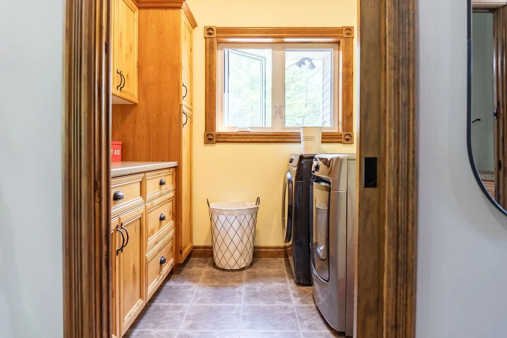 Laundry room with wooden cabinets on the left, a window with wooden frame at the back, a laundry basket, and a washing machine and dryer.