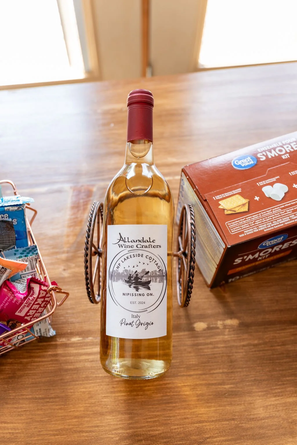 A bottle of white wine labeled 'Aundale Wine Crafters' with a 'Lakeside Cottage' logo, placed on a wooden table. To the right is a box of s'mores ingredients, and to the left are some snacks in a basket.