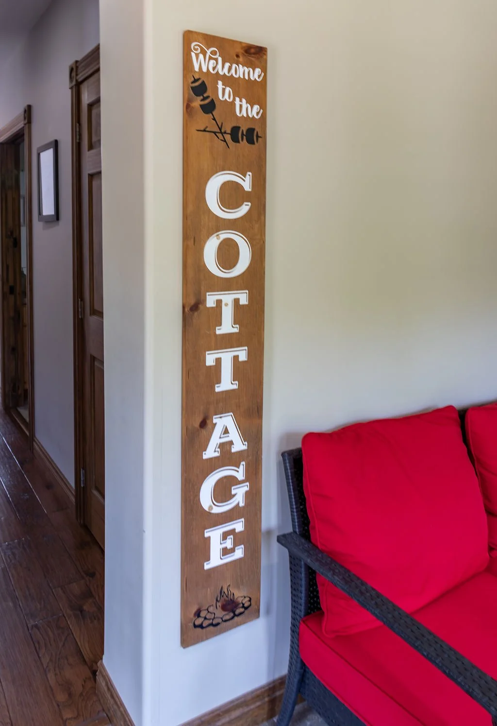 Wooden sign with white letters that say 'Welcome to the Cottage' and a graphic of a skewer with marshmallows and a hot dog, along with a fire pit illustration at the bottom, placed on a corner wall near a red sofa.