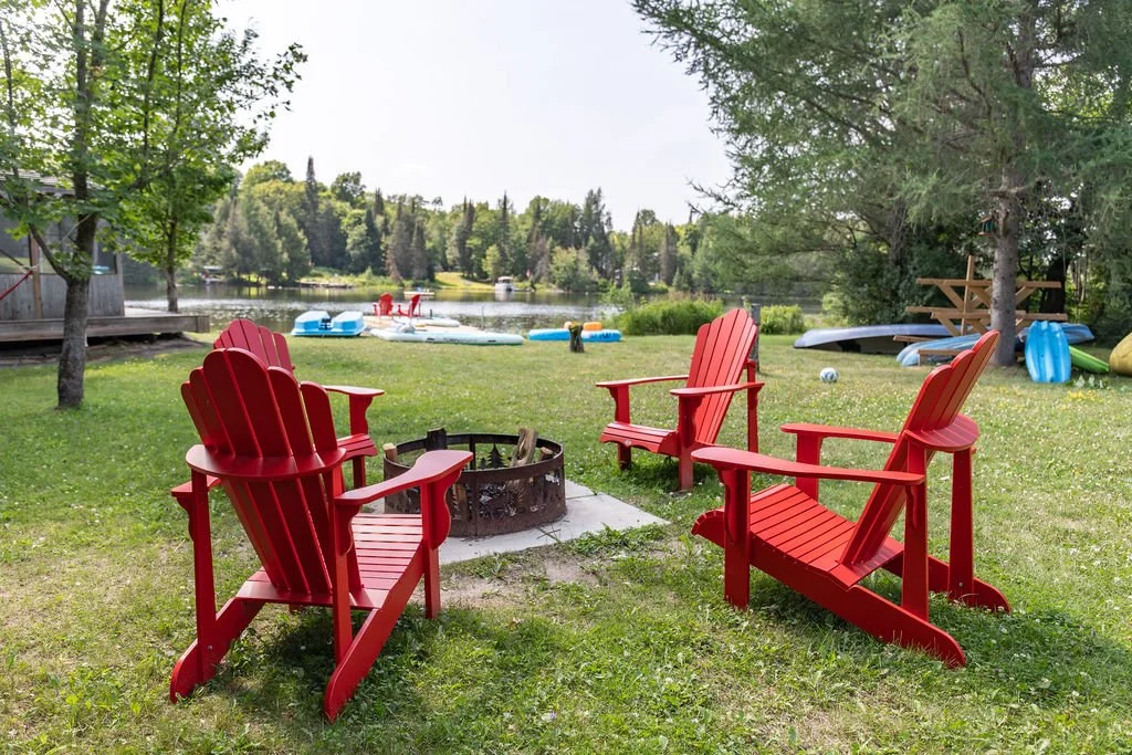 Four red Adirondack chairs arranged in a circle around a fire pit on a grassy area near a lake, with trees and paddle boats in the background.