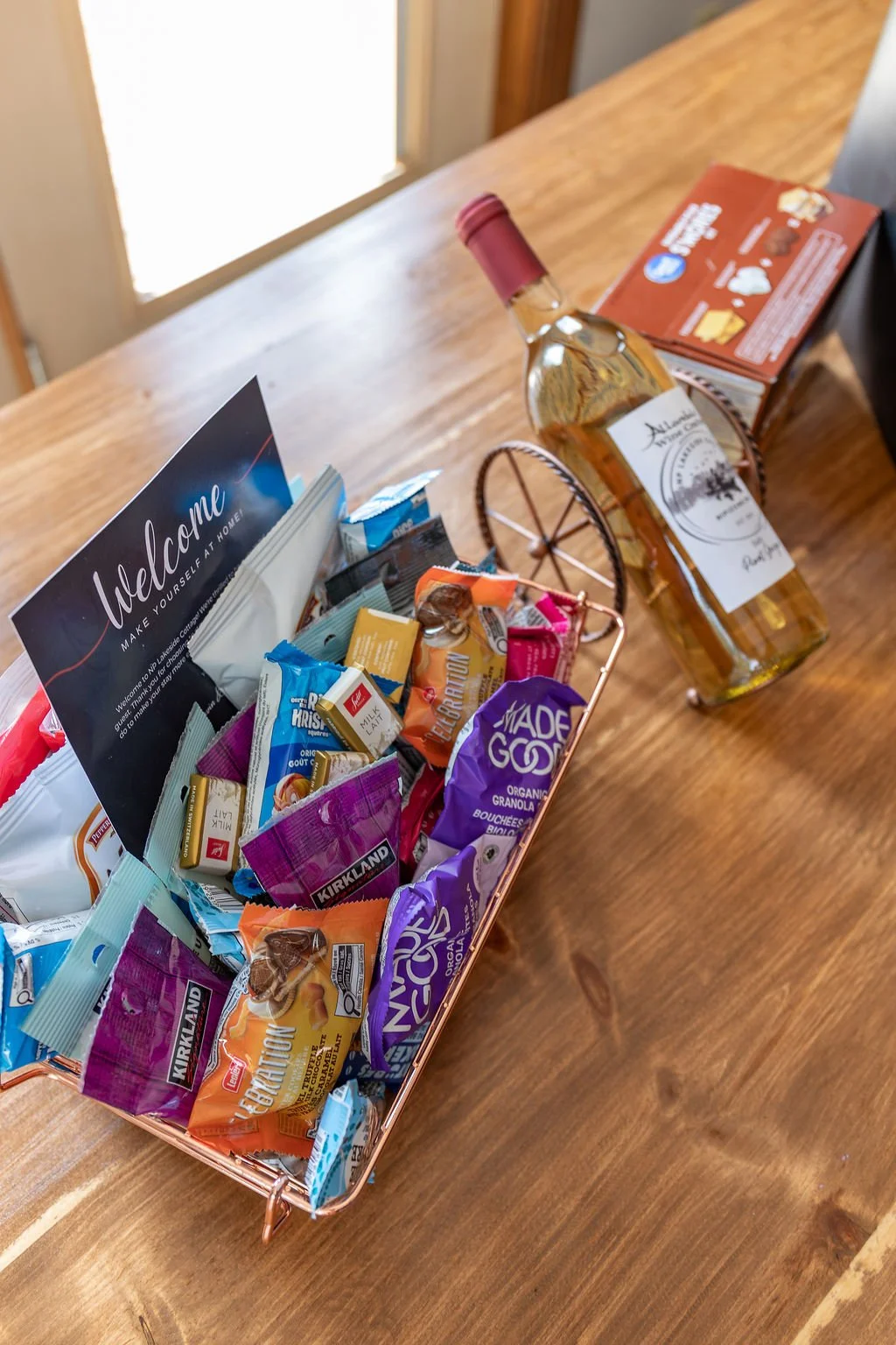 A wooden table with a basket of assorted snacks, a welcome note, and a bottle of white wine in a decorative holder.