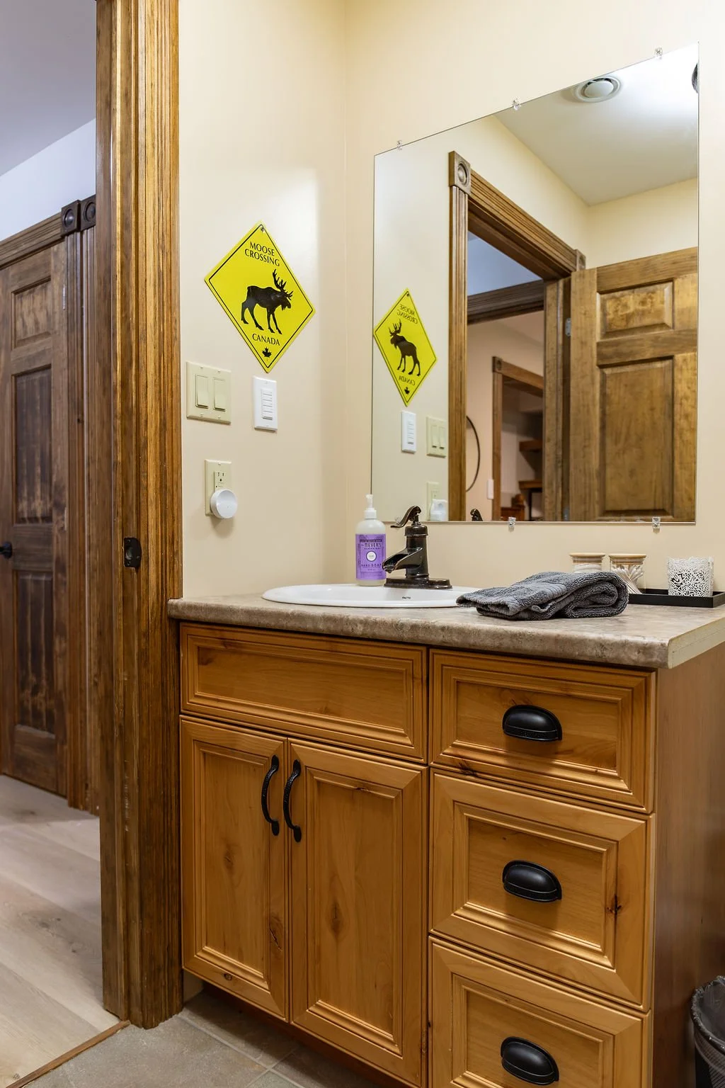 Bathroom with wooden cabinets, a beige countertop, a rectangular mirror, and a black faucet. A hand sanitizer bottle and folded towels are on the counter. Two yellow moose crossing signs are on the wall, and there are wooden doors and trim visible.