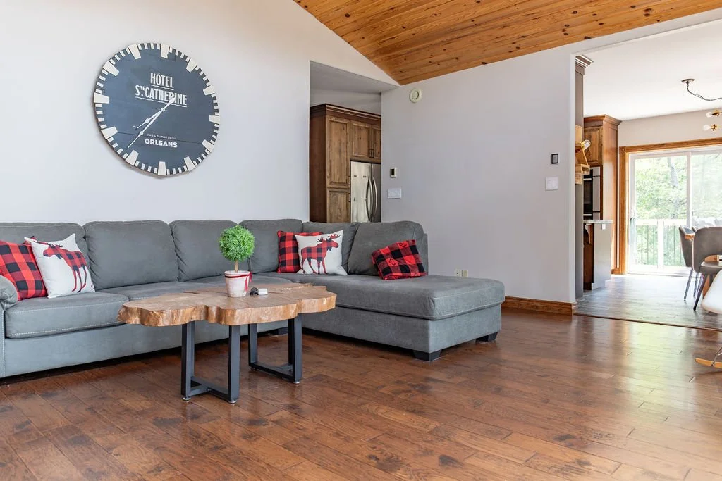 Living room with a gray sectional sofa with decorative pillows resembling reindeer and a buffalo check pattern, a wooden coffee table with a small topiary plant, and a large wall clock indicating it is around 8:12, with a kitchen and sliding glass do