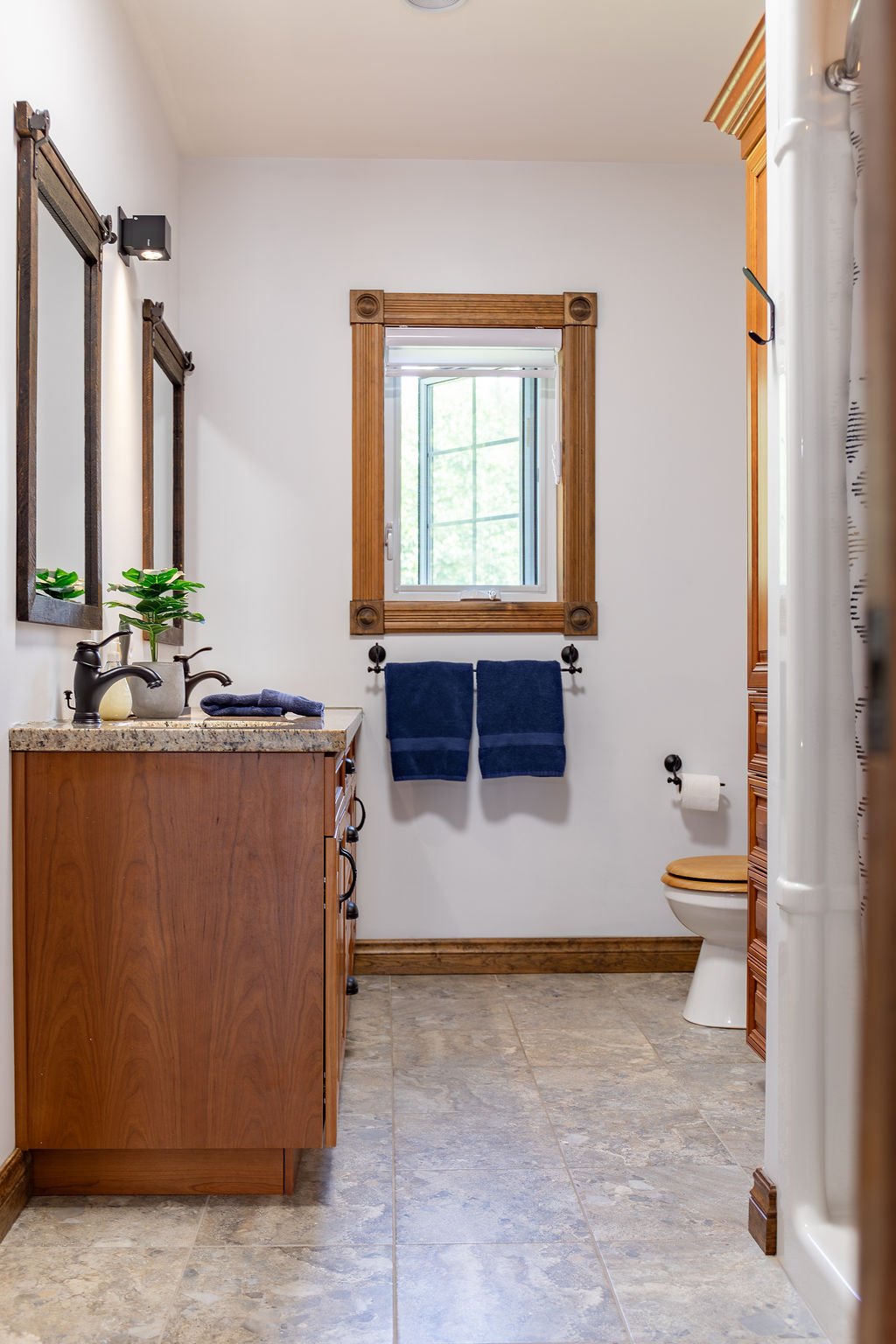Bathroom with wooden accents, a window with wooden frame, a vanity with a sink, a plant, two dark towels on a rod, a toilet with a wooden seat, and a cabinet.