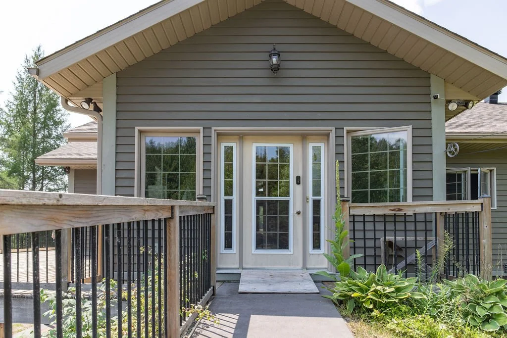 front entrance of a gray house with white trim, featuring a glass-paneled door and two large windows, with a small porch, black metal railing, and garden plants in front.