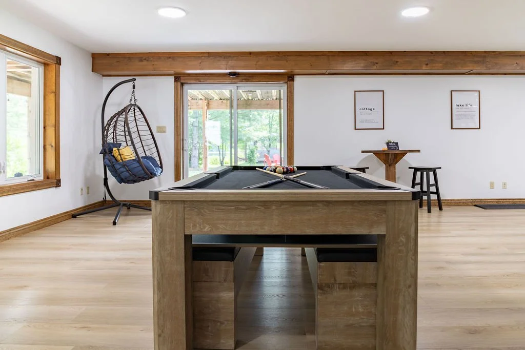A spacious room with light wood flooring, a pool table in the center with pool cues and balls, a hanging egg chair with blue cushions on the left, large windows on the left and sliding glass doors at the back showing a green outdoor area, and framed 