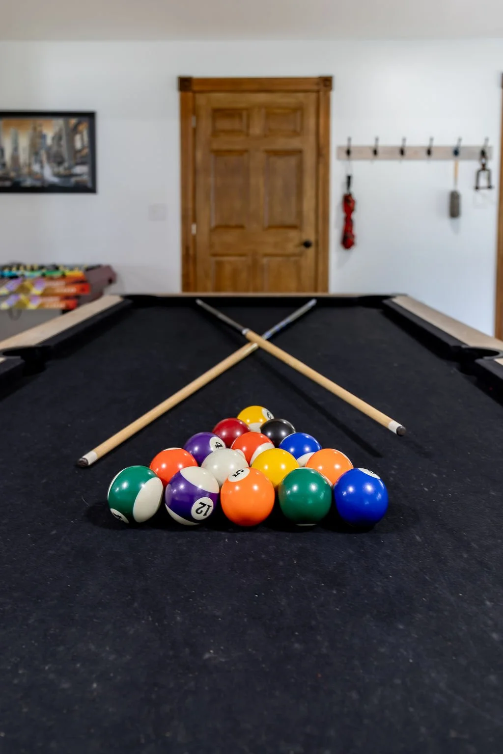 Pool table with billiard balls racked, two cues resting on the table, in a room with a wooden door and wall hooks in the background.