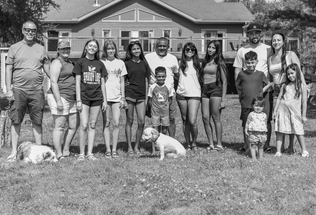 A large group of people, including men, women, children, and dogs, standing on a grassy lawn in front of a house on a sunny day.