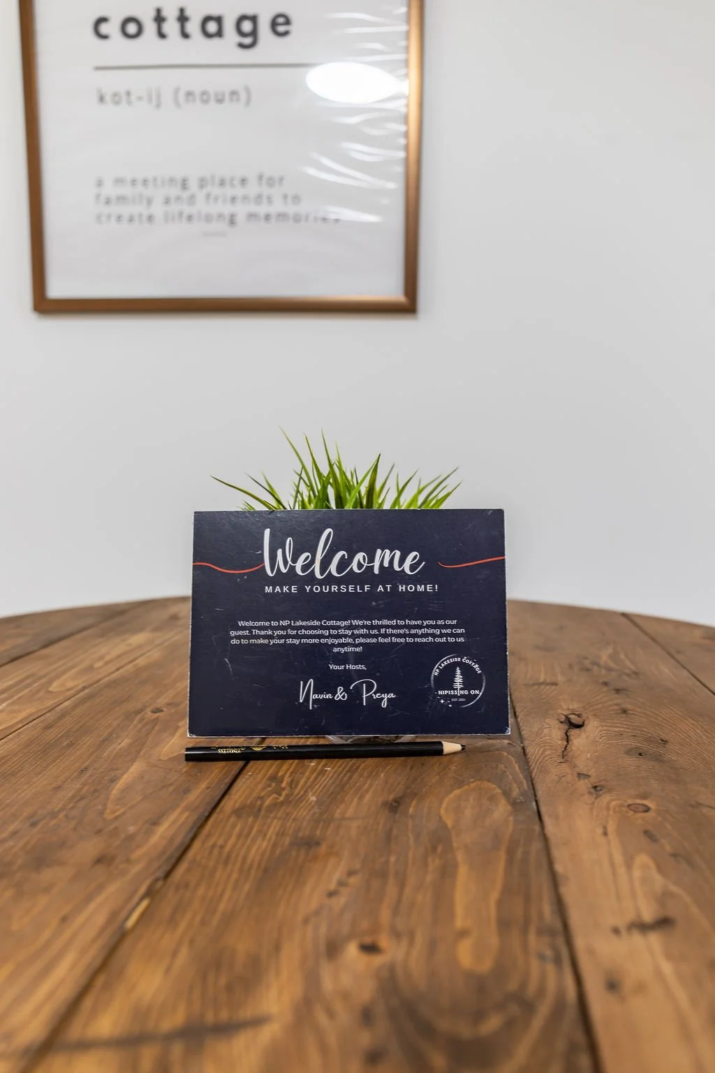A welcome sign on a wooden table with a small green plant in the background and a pen in front. The sign reads 'Welcome, Make Yourself at Home!' and is from NP Lakeside Cottage.
