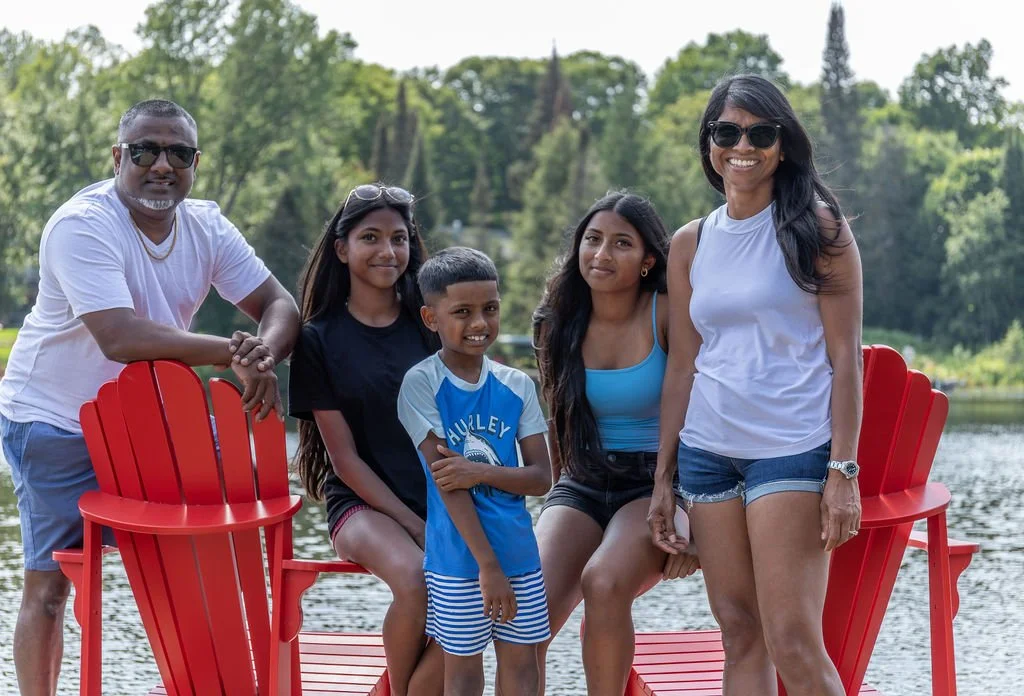 A family of six posing outdoors by a lake with trees in the background. They are smiling and standing around red Adirondack chairs.