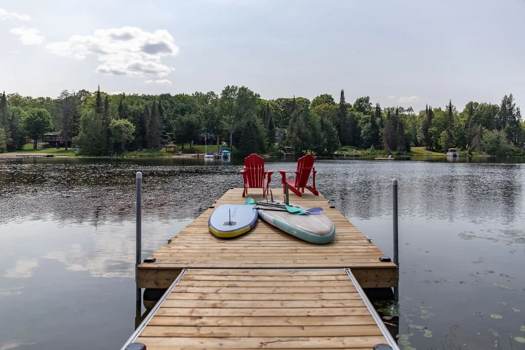 A wooden dock extending into a calm lake with two red Adirondack chairs, a paddleboard, and a paddle resting on it. The background features green trees and a partly cloudy sky.