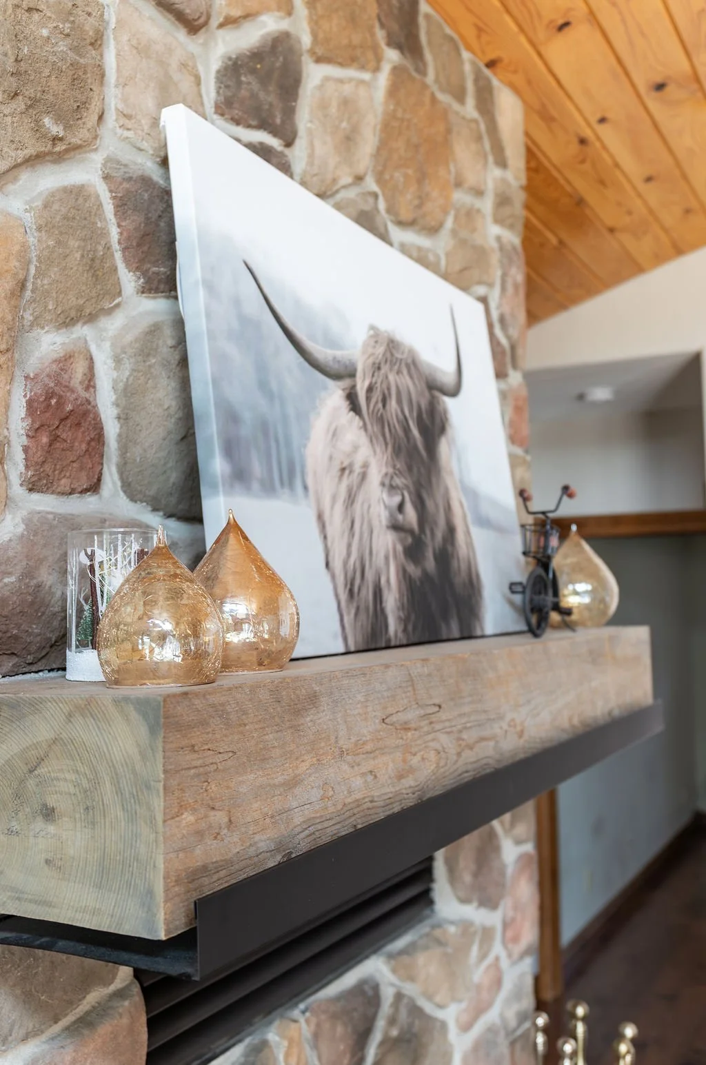 Decorative living room mantle with a framed Highland cow photograph, glass and ceramic vases, and a small vintage bicycle sculpture, all on a wooden mantel against a stone wall with a wooden ceiling.