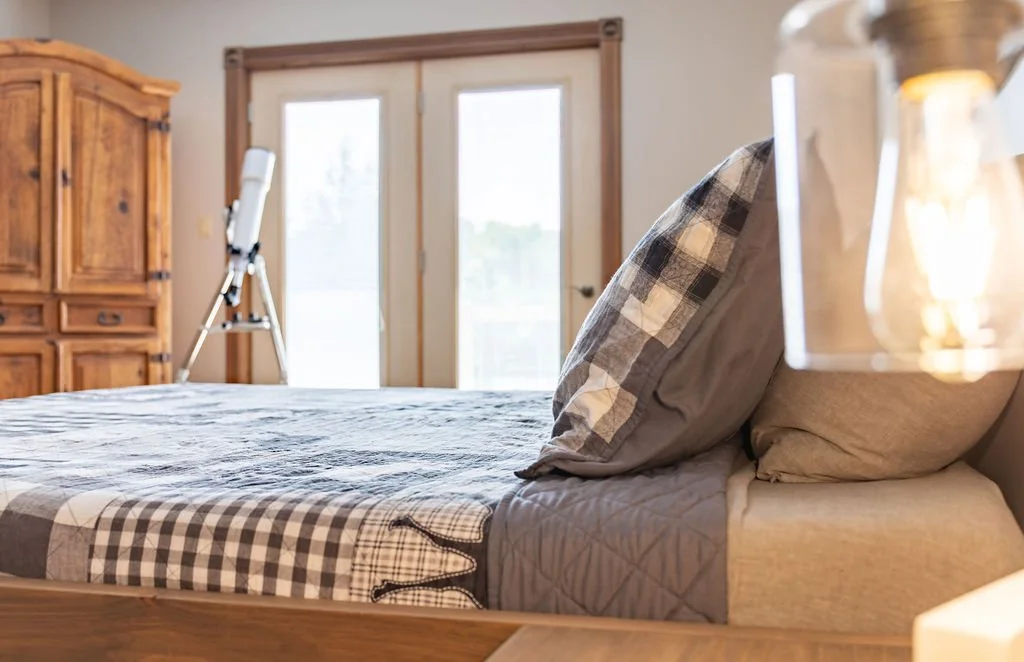 A cozy bedroom with a bed, a checkered blanket, pillows, a wooden wardrobe, and a telescope facing the window.