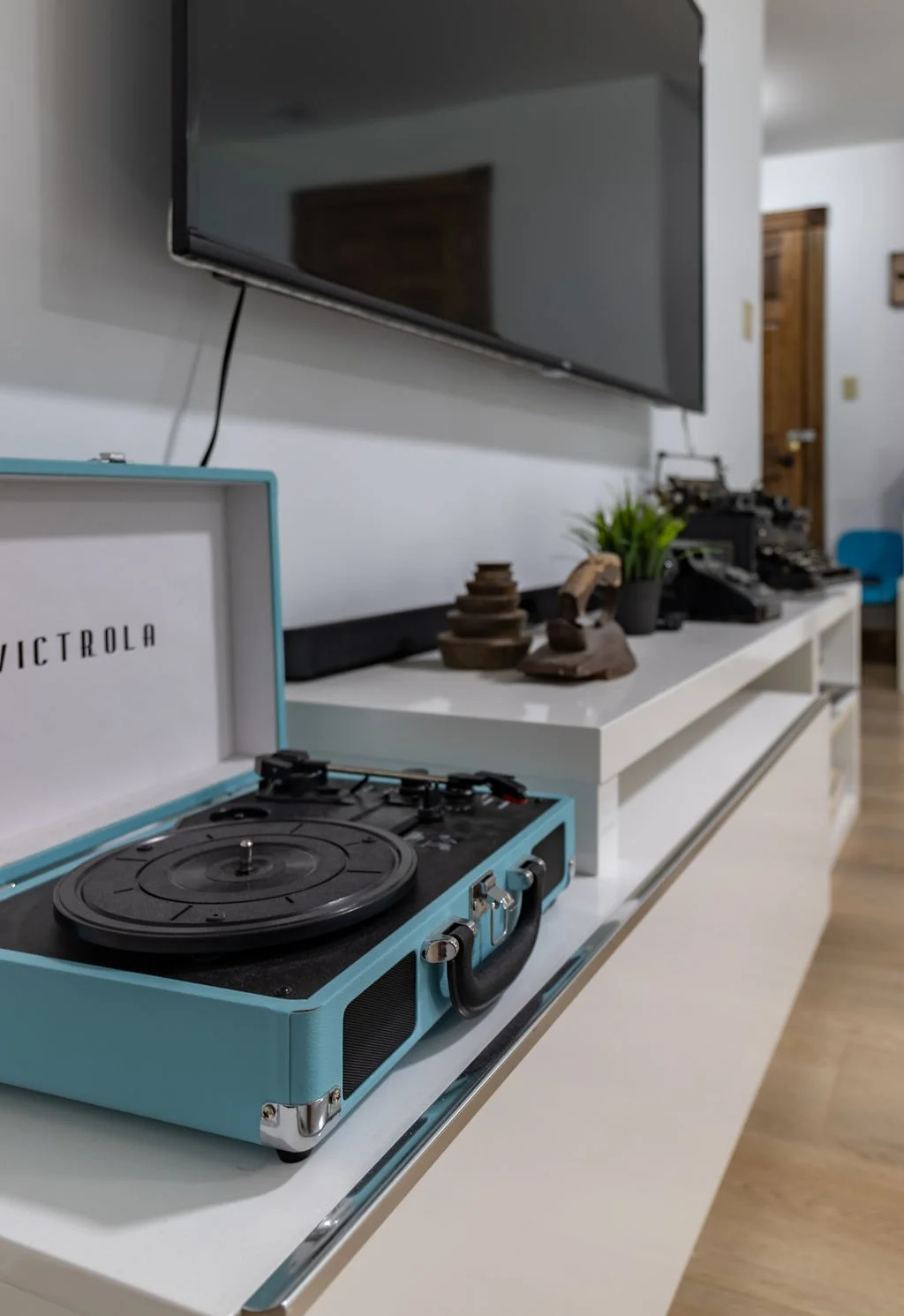 A vintage Victrola record player in a light blue case on a white cabinet with decor, including a plant, a typewriter, and other objects, in a room with wooden doors and flooring.