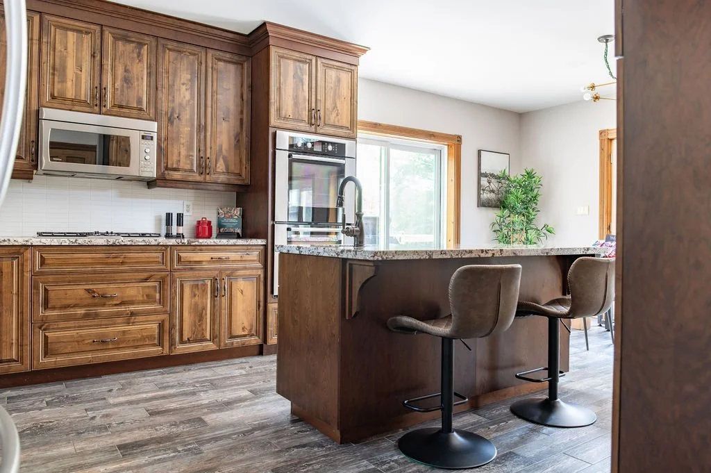 Kitchen with wooden cabinets, granite countertop island with two cushioned bar stools, built-in microwave, oven, gas stove, white tiled backsplash, window showing outdoor scenery, potted plant, and dining area in the background.