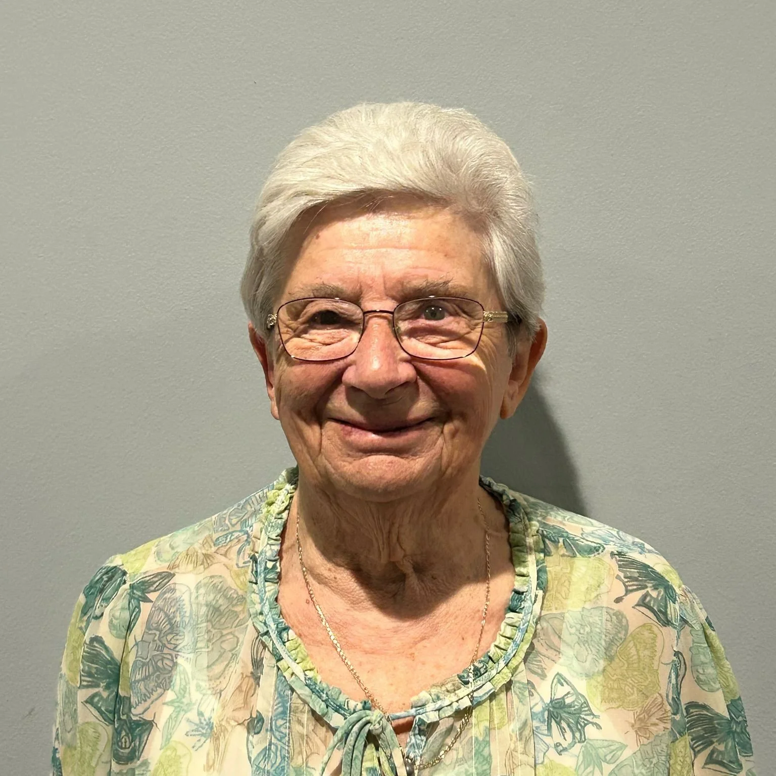 An elderly woman with short white hair and glasses smiling, wearing a floral blouse with a tie at the neckline, standing against a plain gray wall.