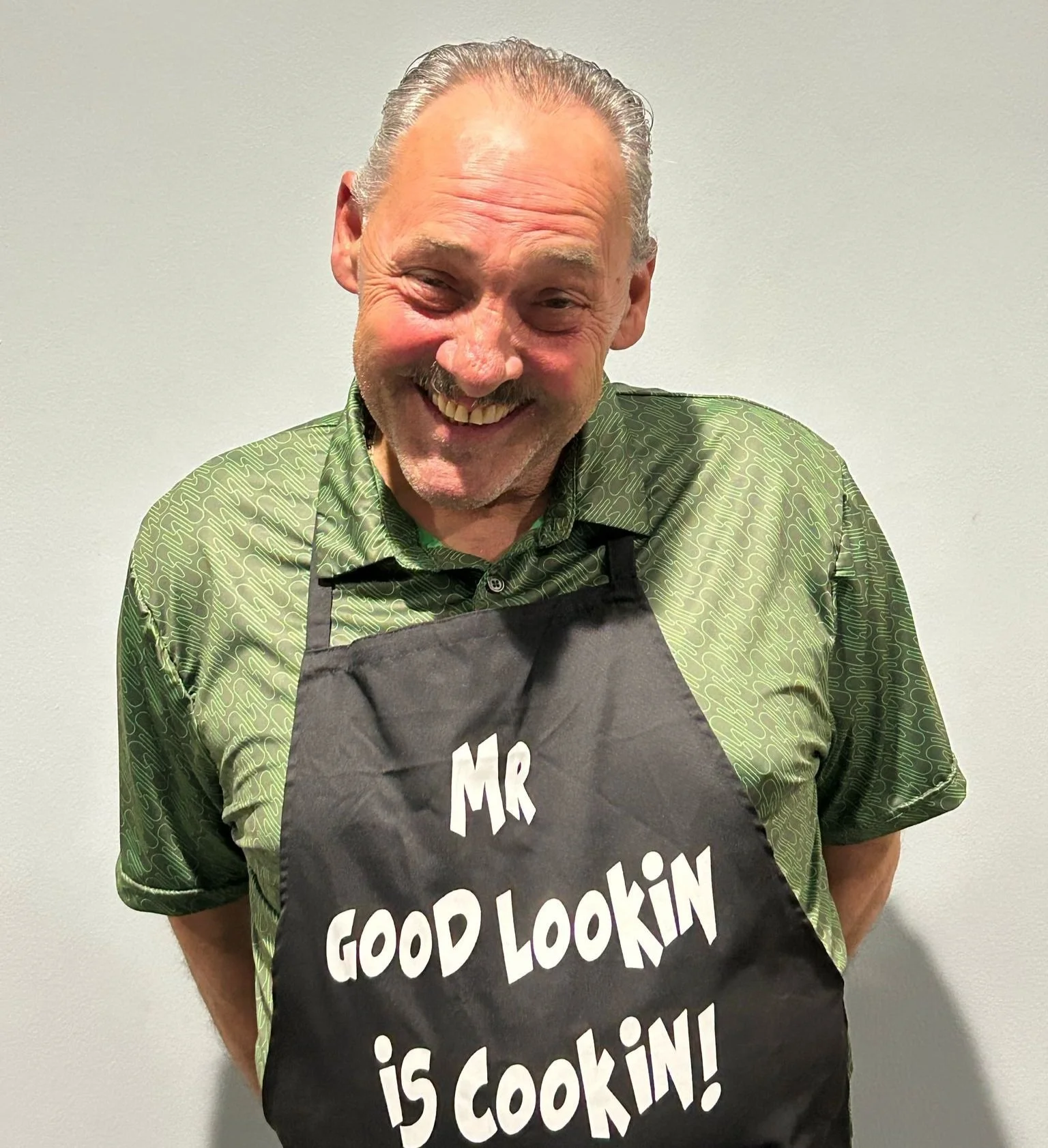 A smiling man with gray hair and a goatee wearing a green patterned shirt and a black apron that says 'Mr. Good Lookin is Cookin!'