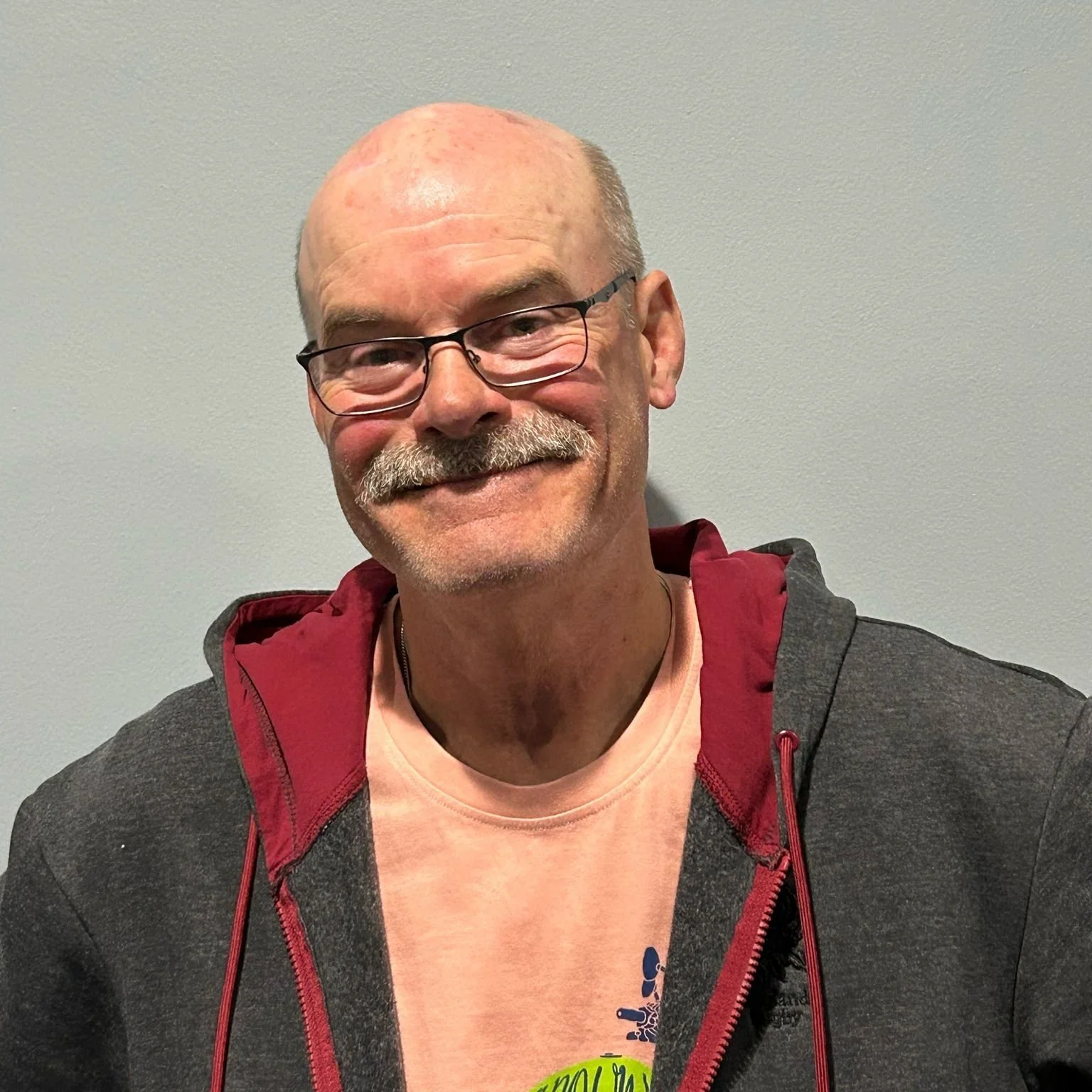 A smiling middle-aged man with glasses, sporting a mustache and beard, wearing a beige T-shirt and a dark zip-up hoodie with red lining, standing against a plain light-colored wall.