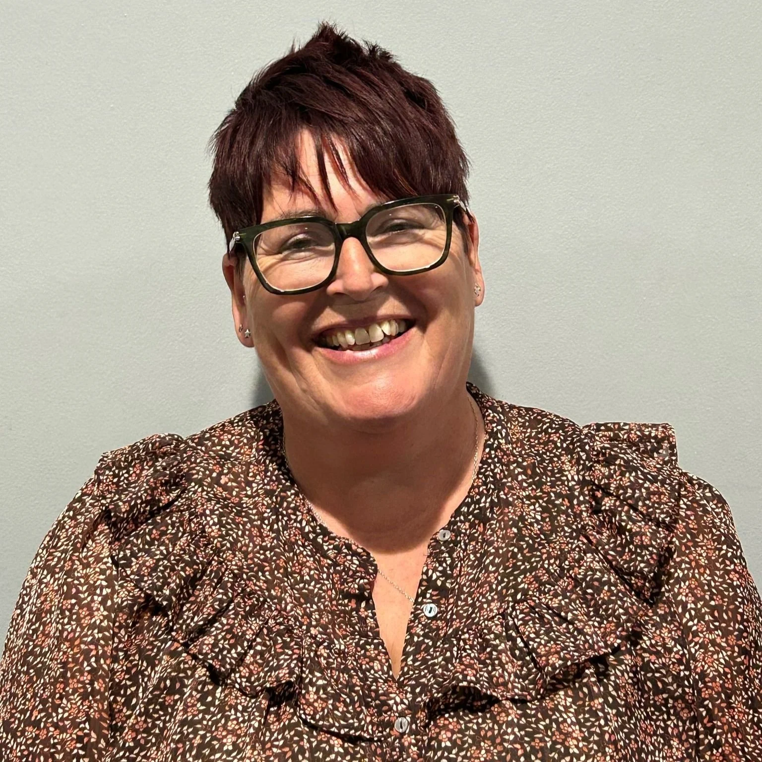 Smiling woman with short red hair, glasses, earrings, and patterned blouse standing against plain light-colored wall.