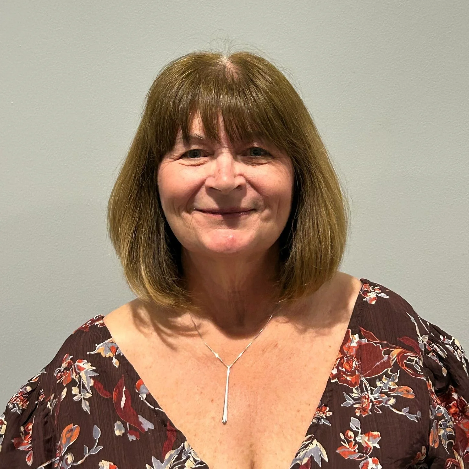 A woman with shoulder-length brown hair, smiling, wearing a floral dress and a delicate necklace with a vertical pendant, standing against a plain light-colored wall.