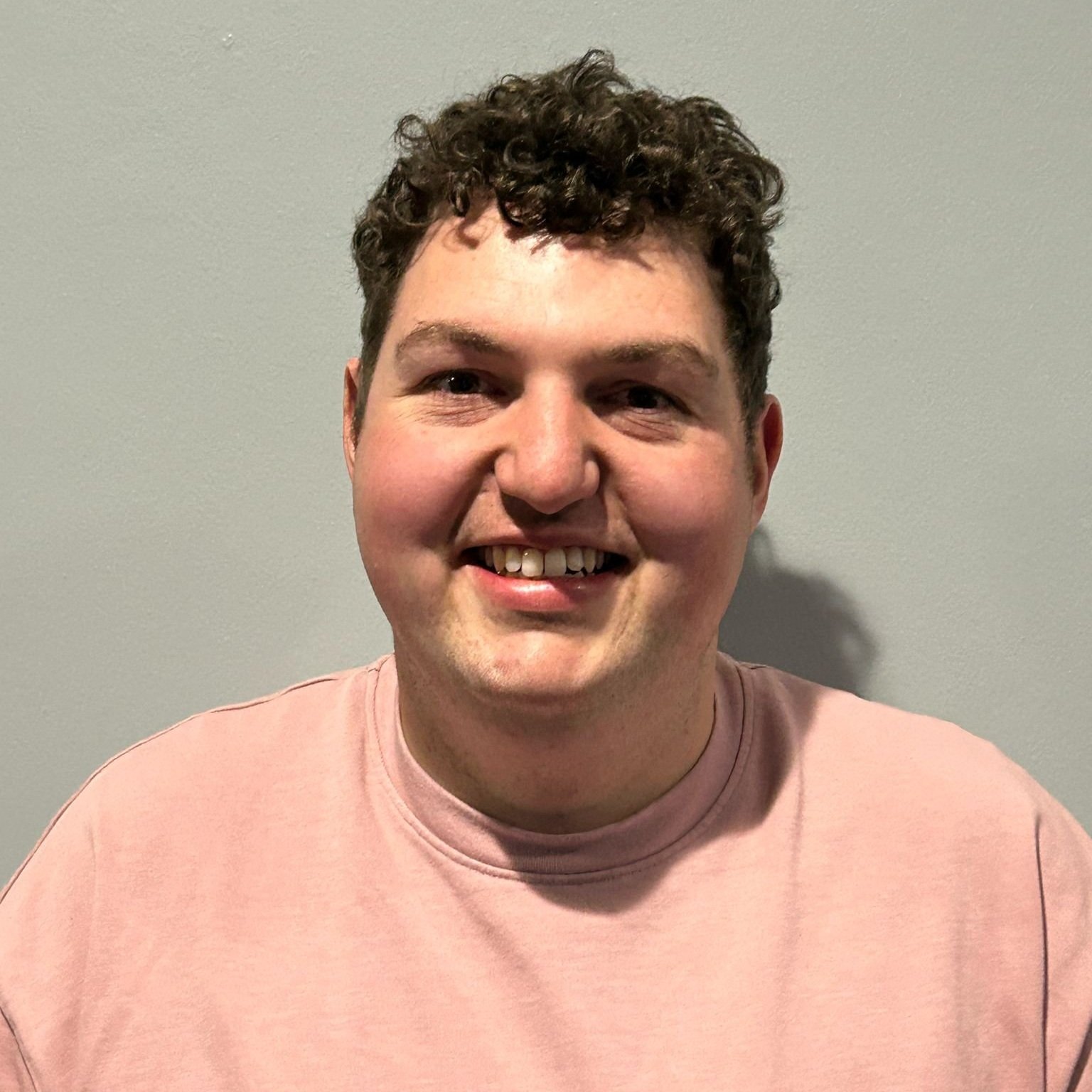 Portrait of a young man with curly brown hair smiling, wearing a light pink shirt, standing against a plain light grey wall.