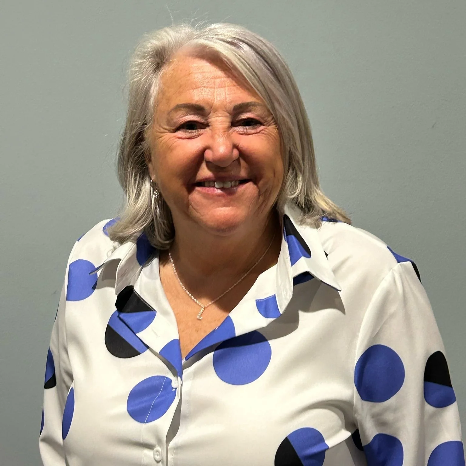 A smiling woman with shoulder-length gray hair wearing a white blouse with blue and black polka dots, standing against a plain gray wall.