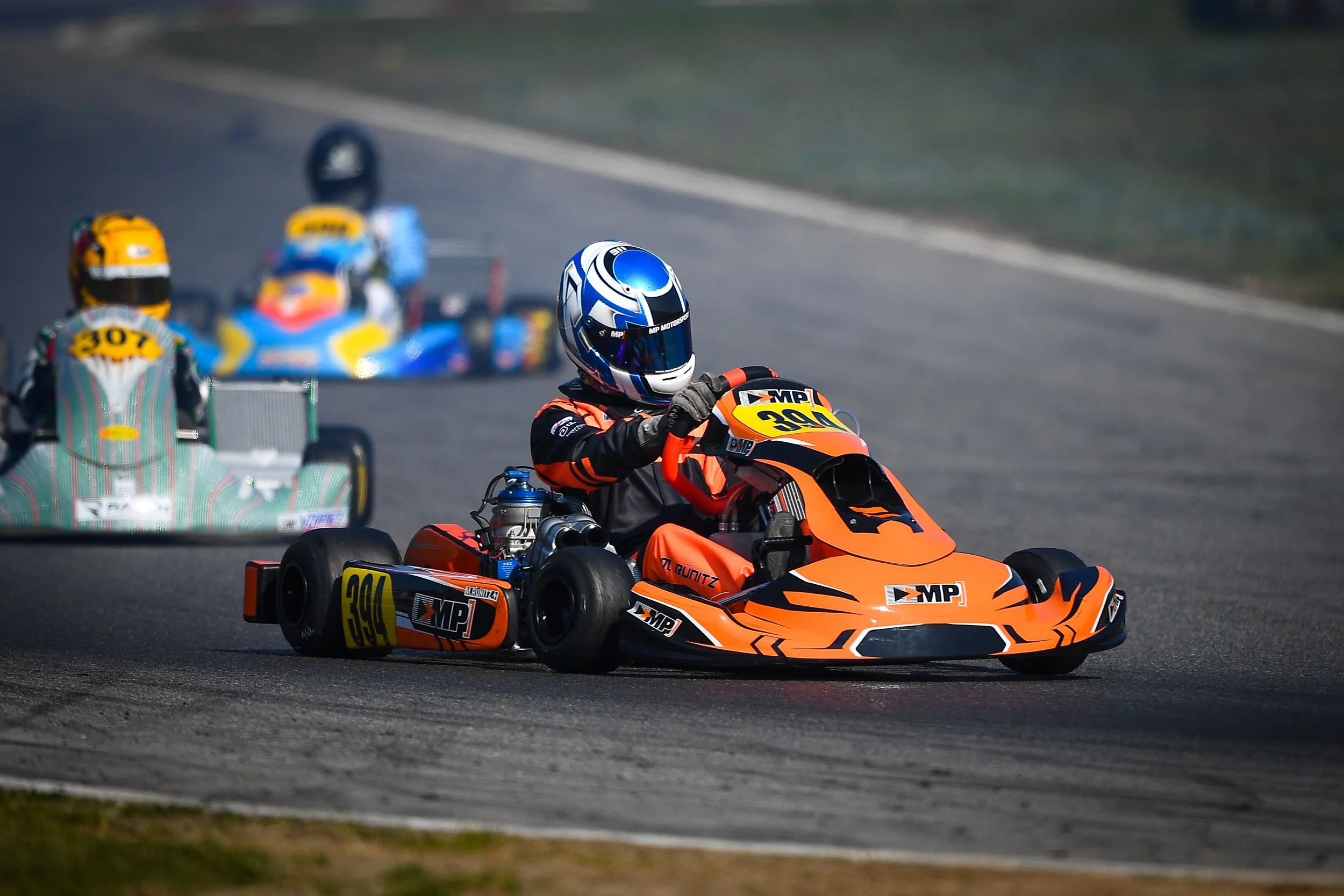 A go-kart race on a track with a driver in an orange and black suit and helmet, leading behind a kart with a driver in a green suit, and a blue kart with a driver in a yellow helmet in the background.