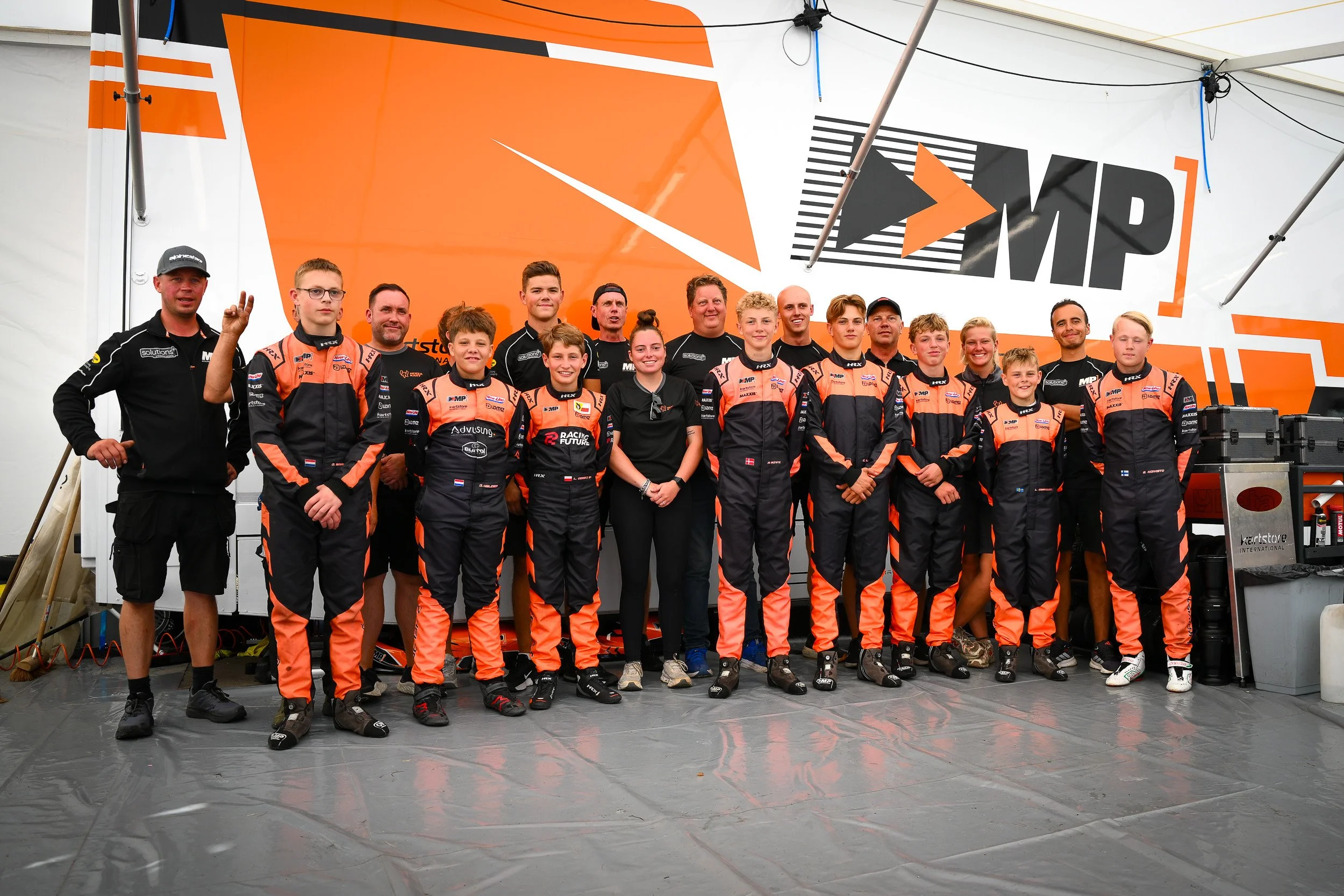 A racing team of 15 members, including drivers in black and orange racing suits and staff, posed indoors under a large orange and white banner with the logo 'MP' and an arrow, all smiling for a team photo.