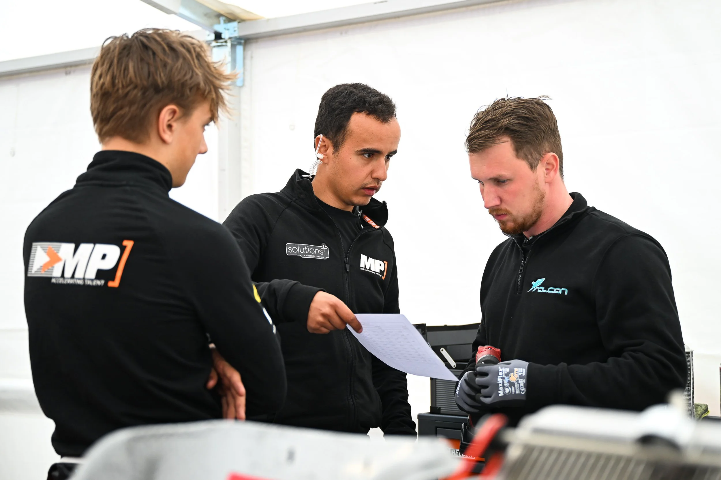 Three men in black jackets discussing a printed document in a white tent.