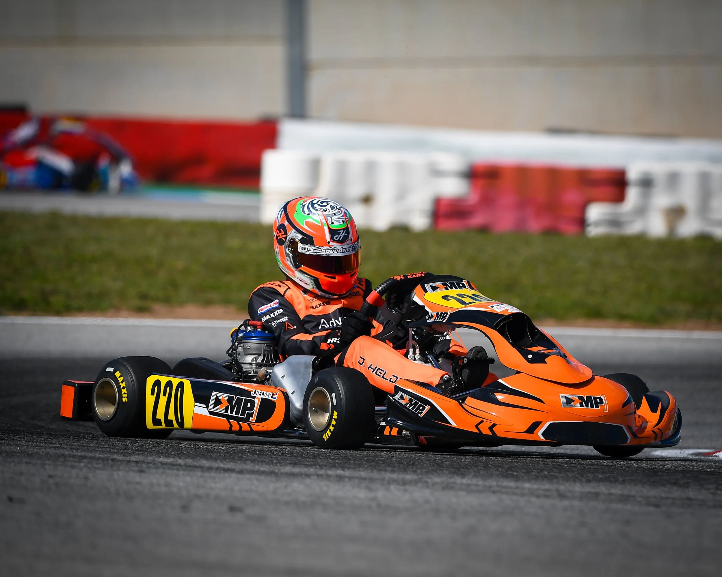 A go-kart racer in orange and black racing gear and helmet driving a bright orange go-kart on a racetrack.