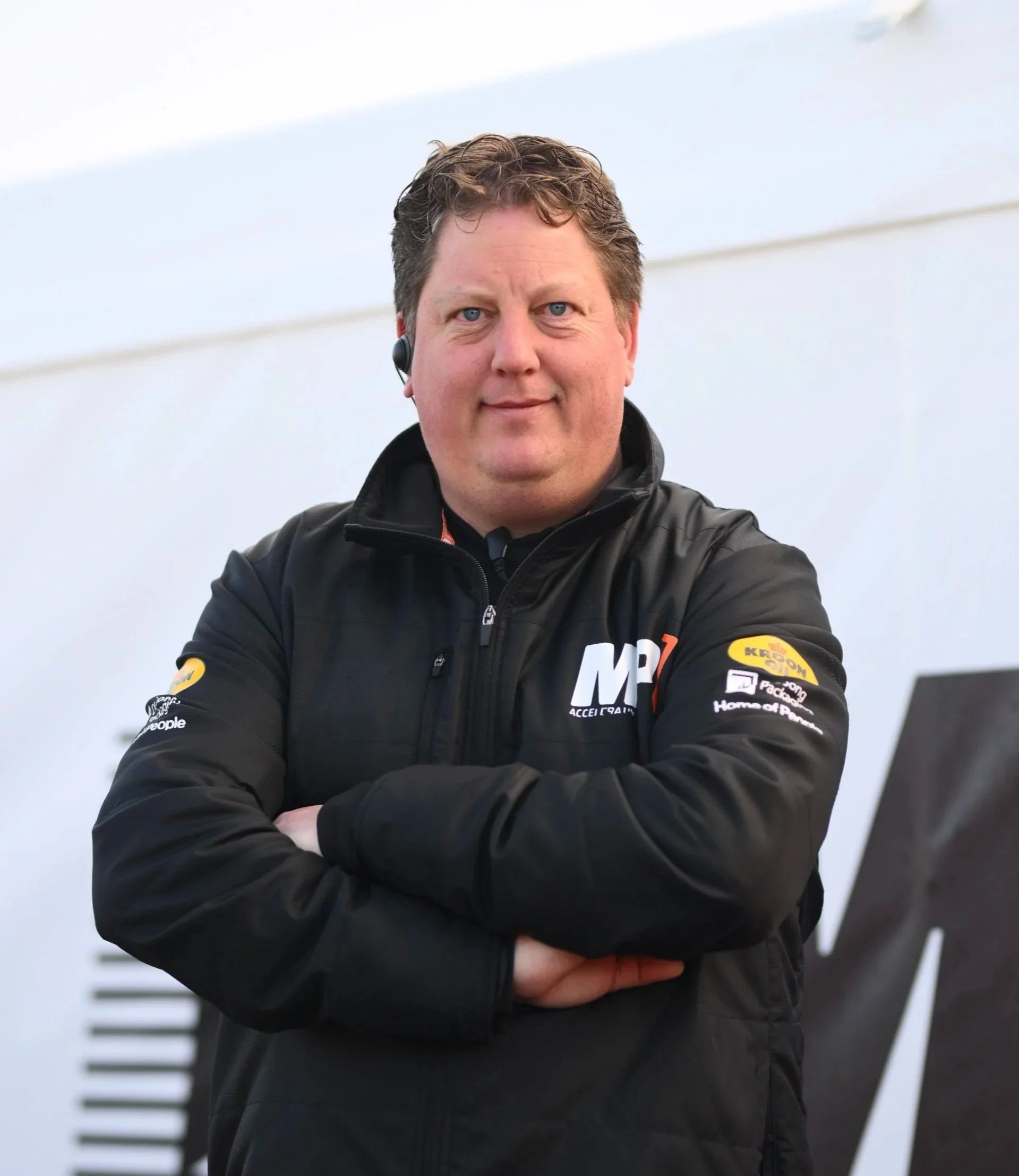 Man standing inside a racing garage next to a racing go-kart, wearing a black jacket with sponsor logos.