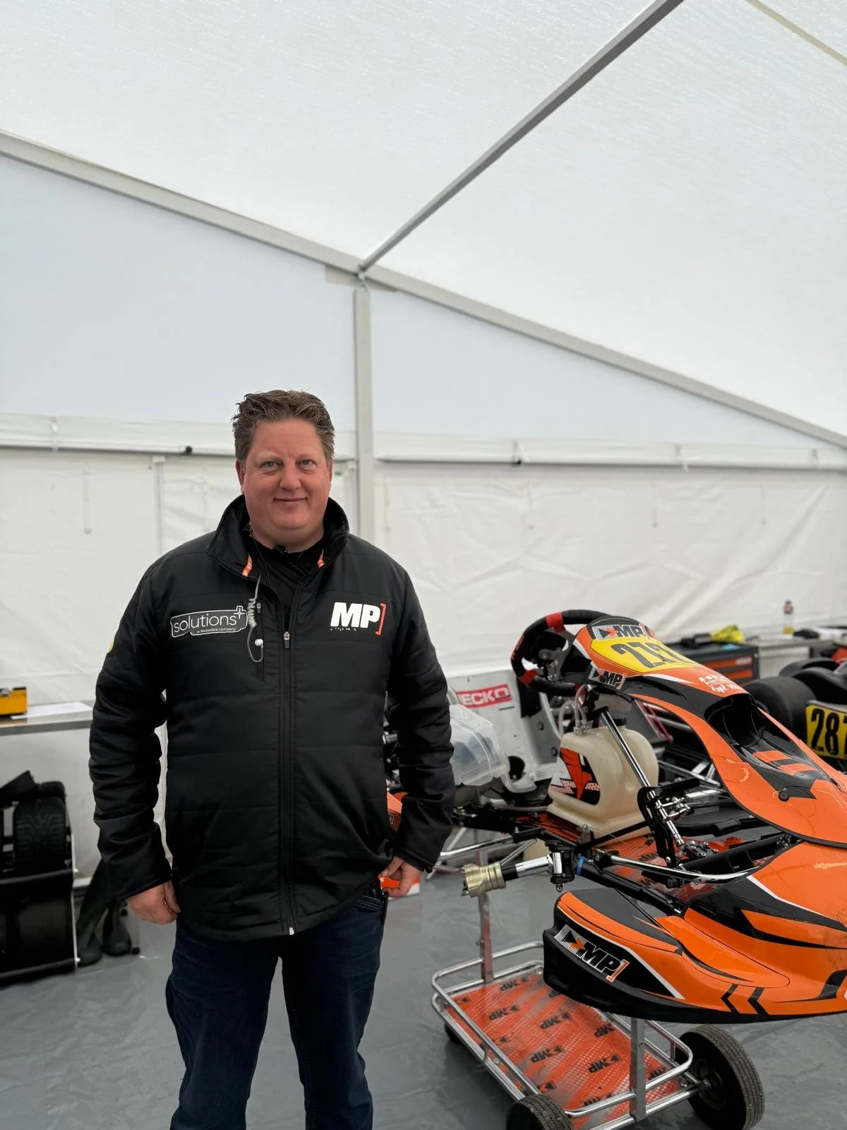 Man standing inside a racing garage next to a racing go-kart, wearing a black jacket with sponsor logos.
