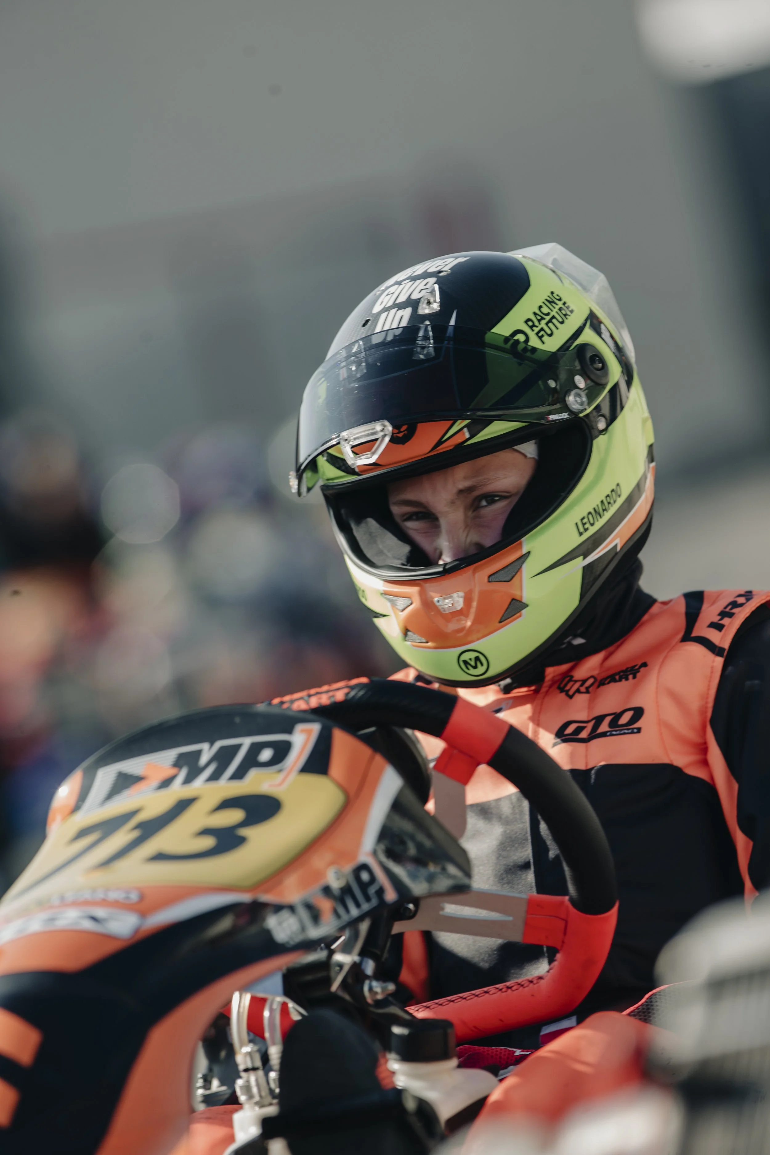 Close-up of a young race car driver in a bright helmet with a clear visor, sitting on a motorcycle with the number 713. The driver is wearing a red and black racing suit with sponsor logos, looking focused before a race.