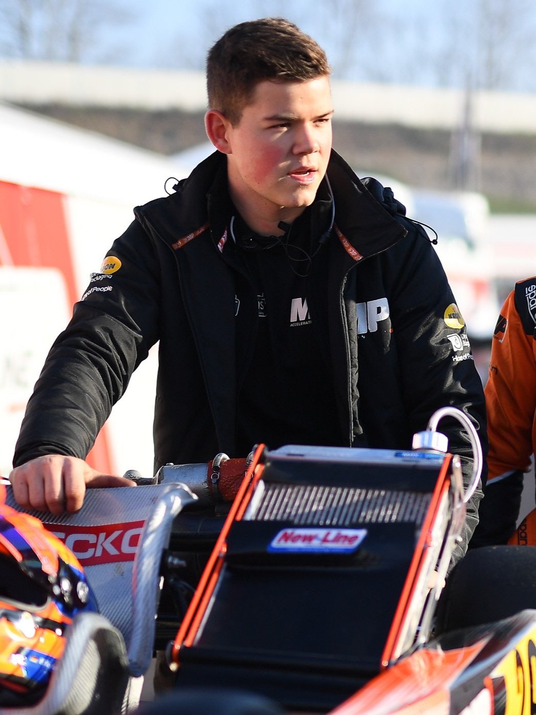 A young man with short brown hair, wearing a black racing jacket with various sponsor logos, stands next to a motorbike during a racing event.