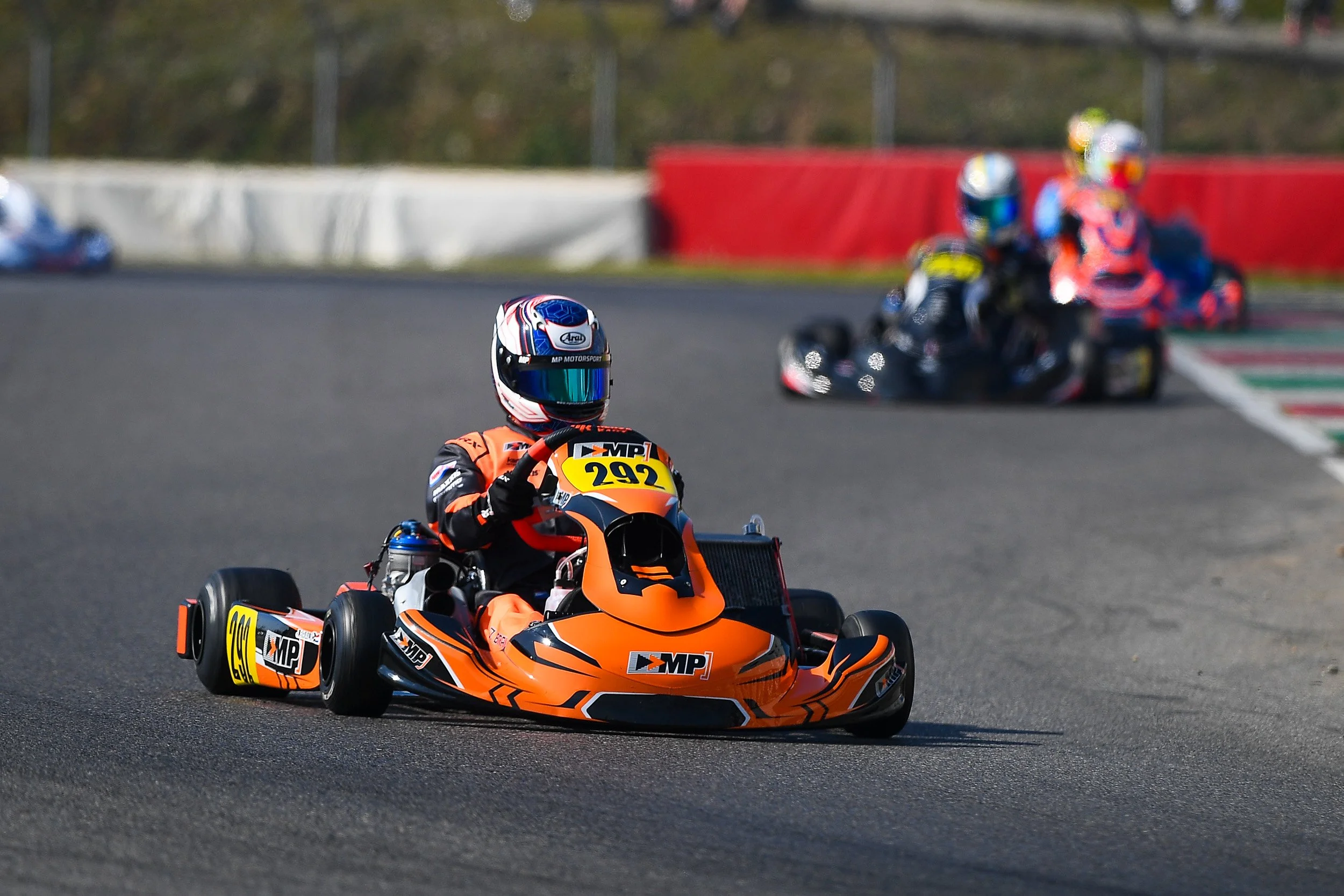 Kart racers on a race track, with one in the foreground and others in the background.