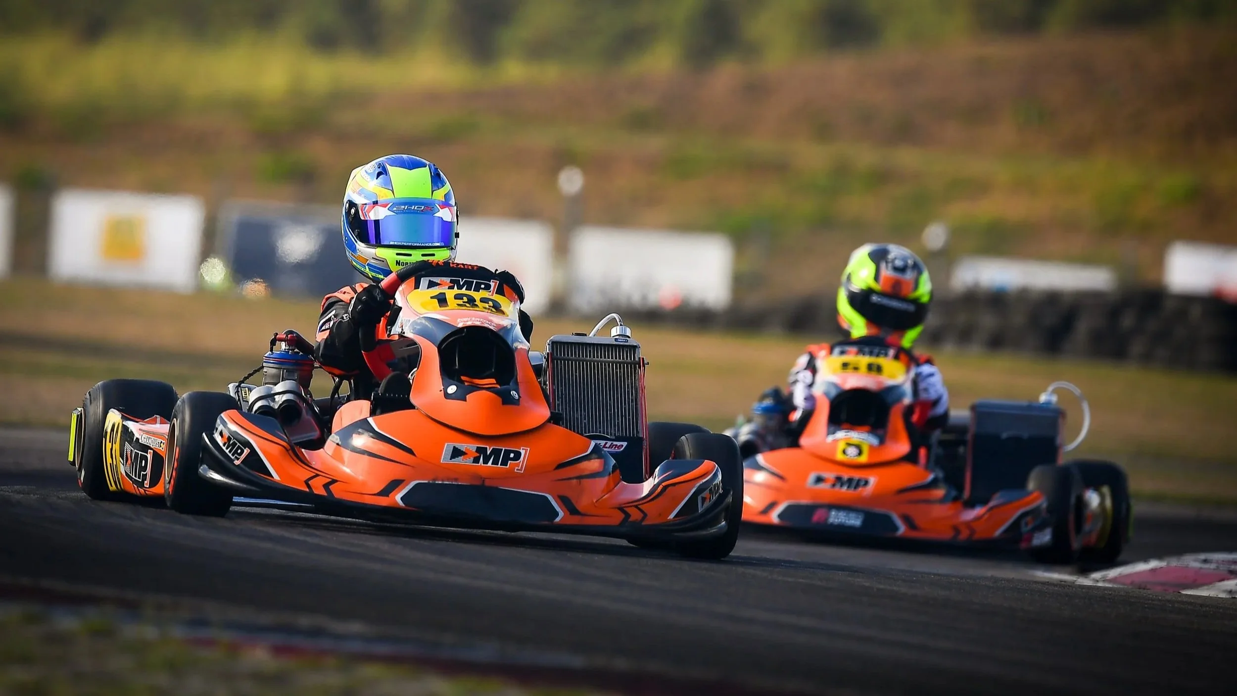 Two go-karts racing on a track, with drivers wearing helmets and racing suits, surrounded by a blurred background of grassy areas and trees.