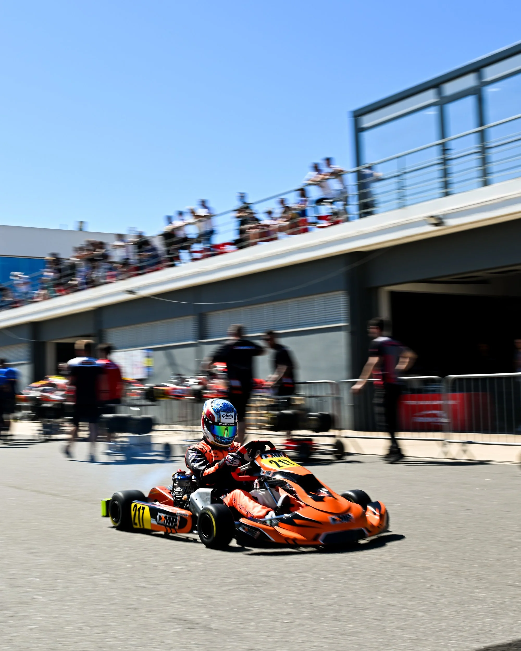 Open-wheel race car in orange and black racing on track, driver wearing a blue helmet, pit crew and spectators in the background.