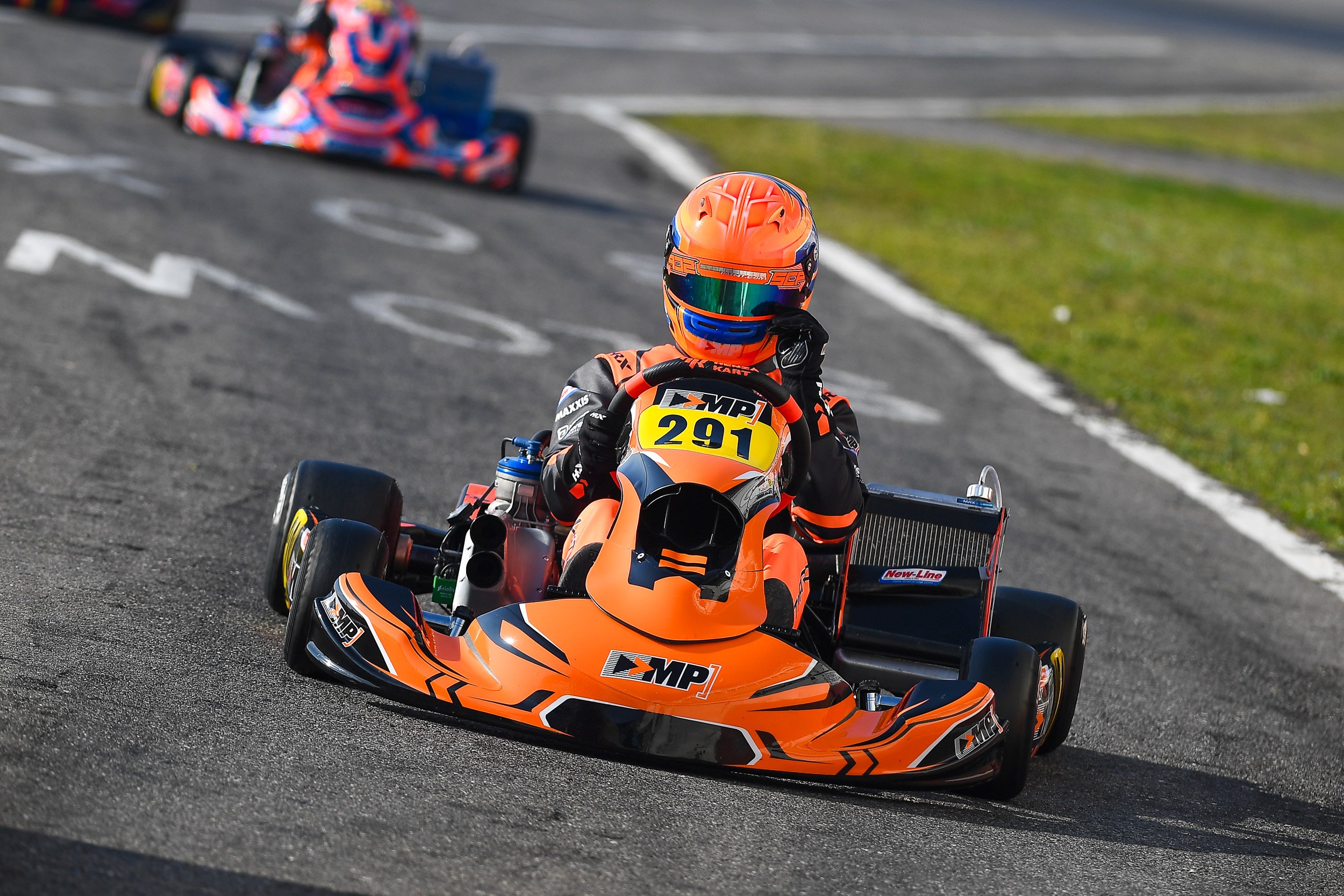 A go-kart driver in orange racing gear and helmet racing on a track, with another go-kart visible in the background.