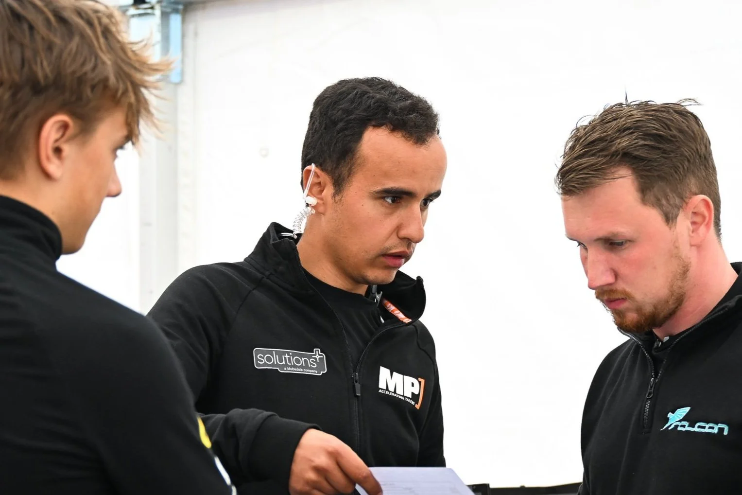 Three men in black jackets looking at a document, one of them with a headset, in a white background setting.