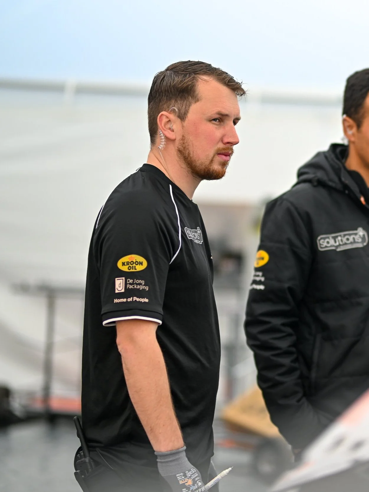 A man with short brown hair, beard, and earpiece, wearing a black shirt with sponsor logos, stands outdoors with a serious expression.