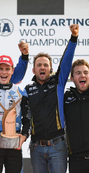 Three men celebrating on a podium with a FIA Karting World Championship backdrop, one holding a trophy.