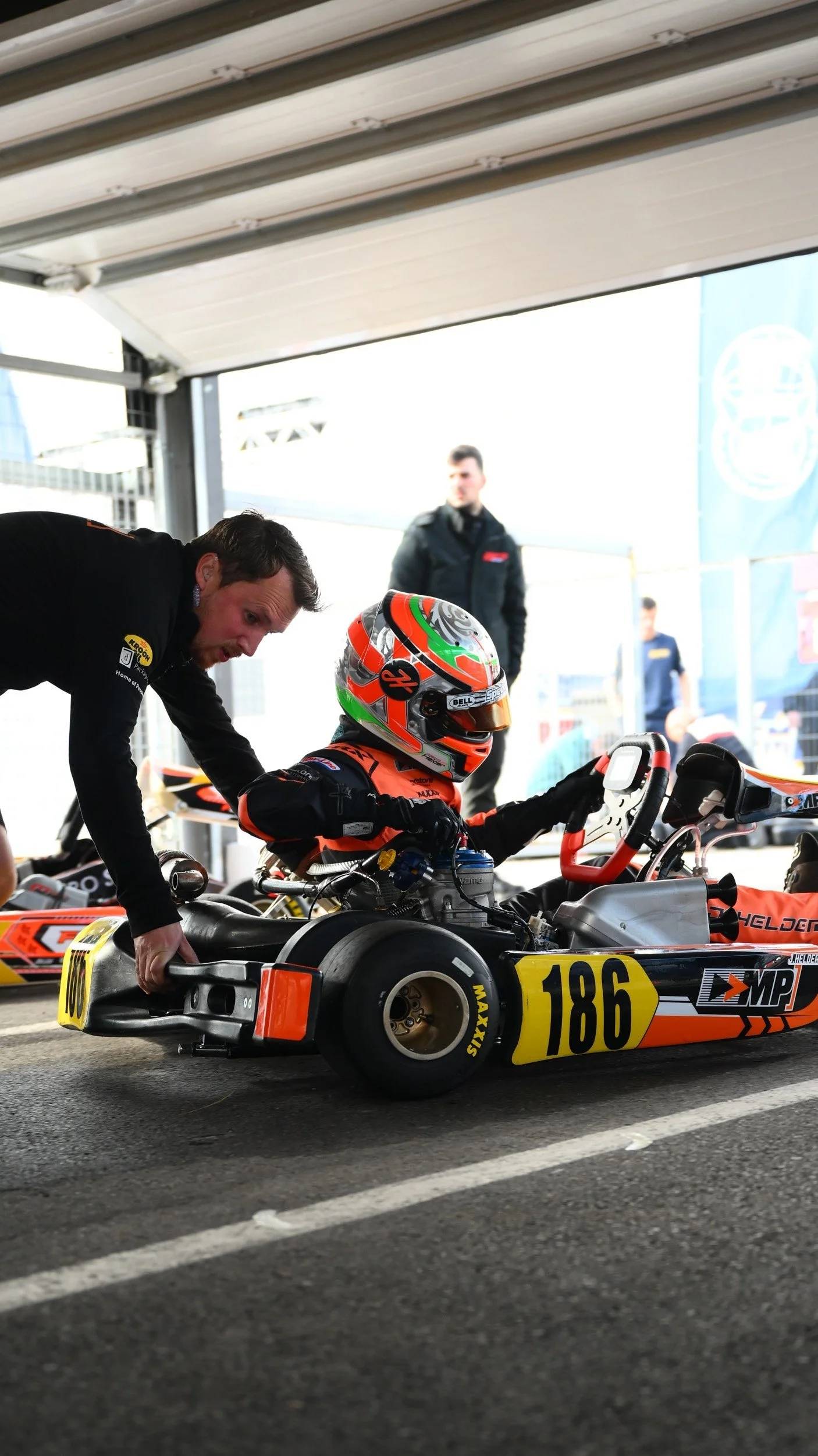 A race car driver in a racing suit and helmet sitting in a go-kart under a shelter, with mechanics working on it and other people in the background.