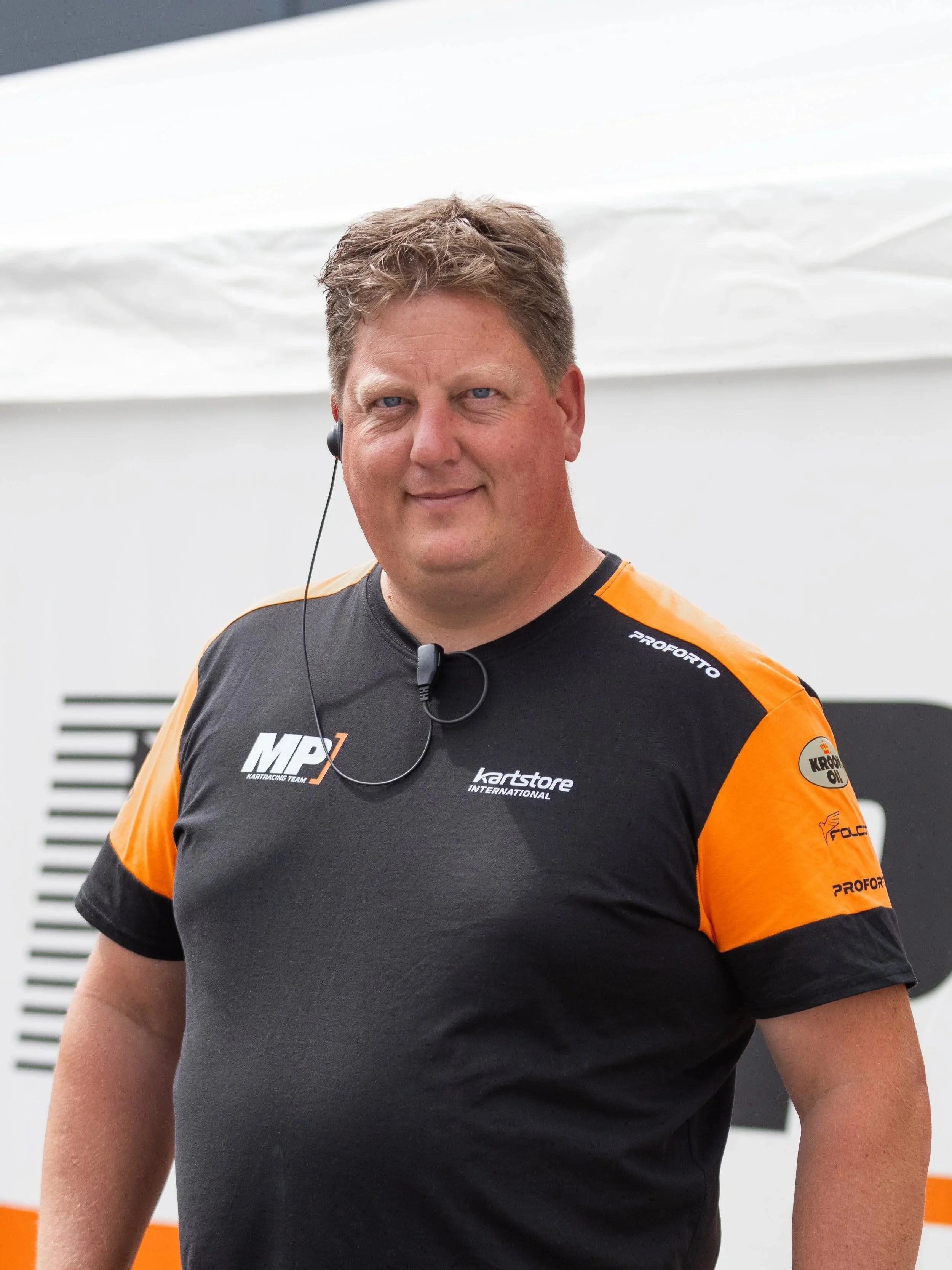 Man standing inside a racing garage next to a racing go-kart, wearing a black jacket with sponsor logos.