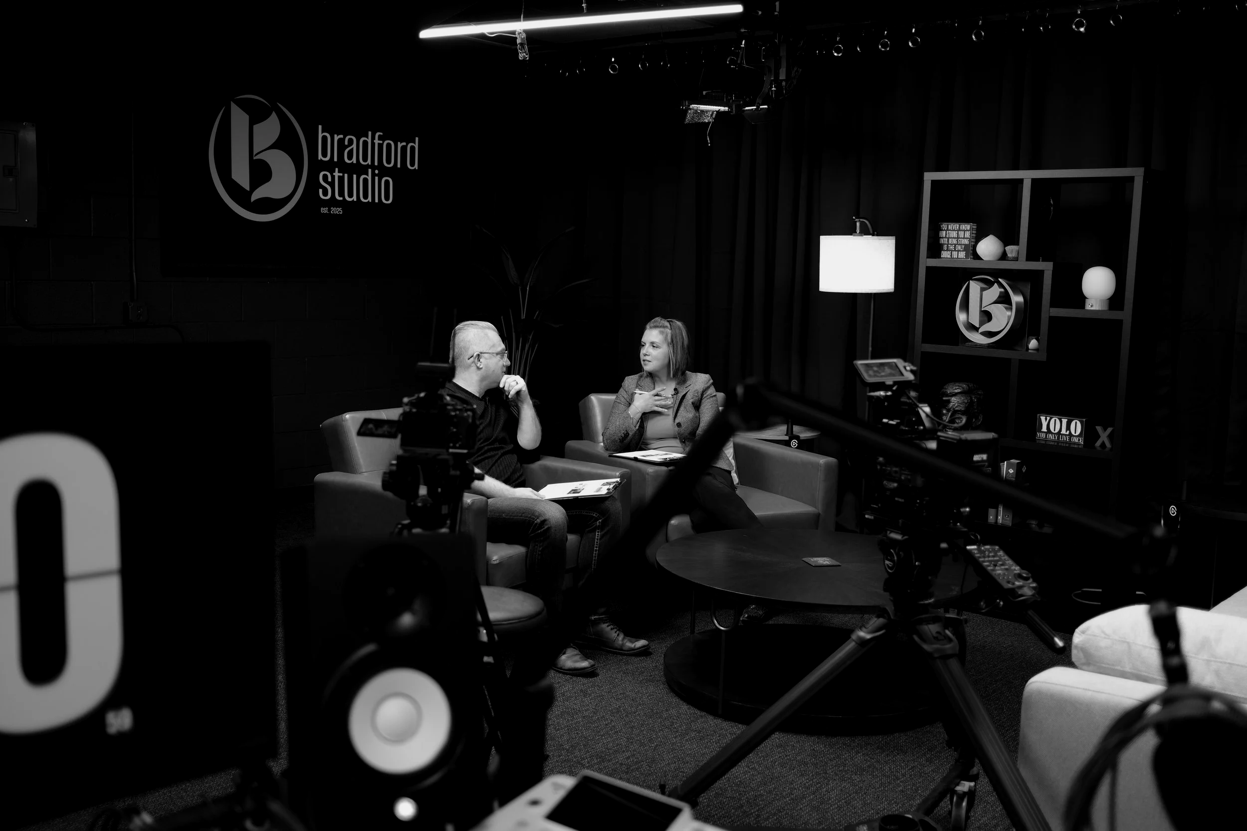 Two people sitting on a sofa having a conversation in a recording studio, with cameras and lighting equipment in the foreground and a logo on the wall that reads 'bradford studio'.