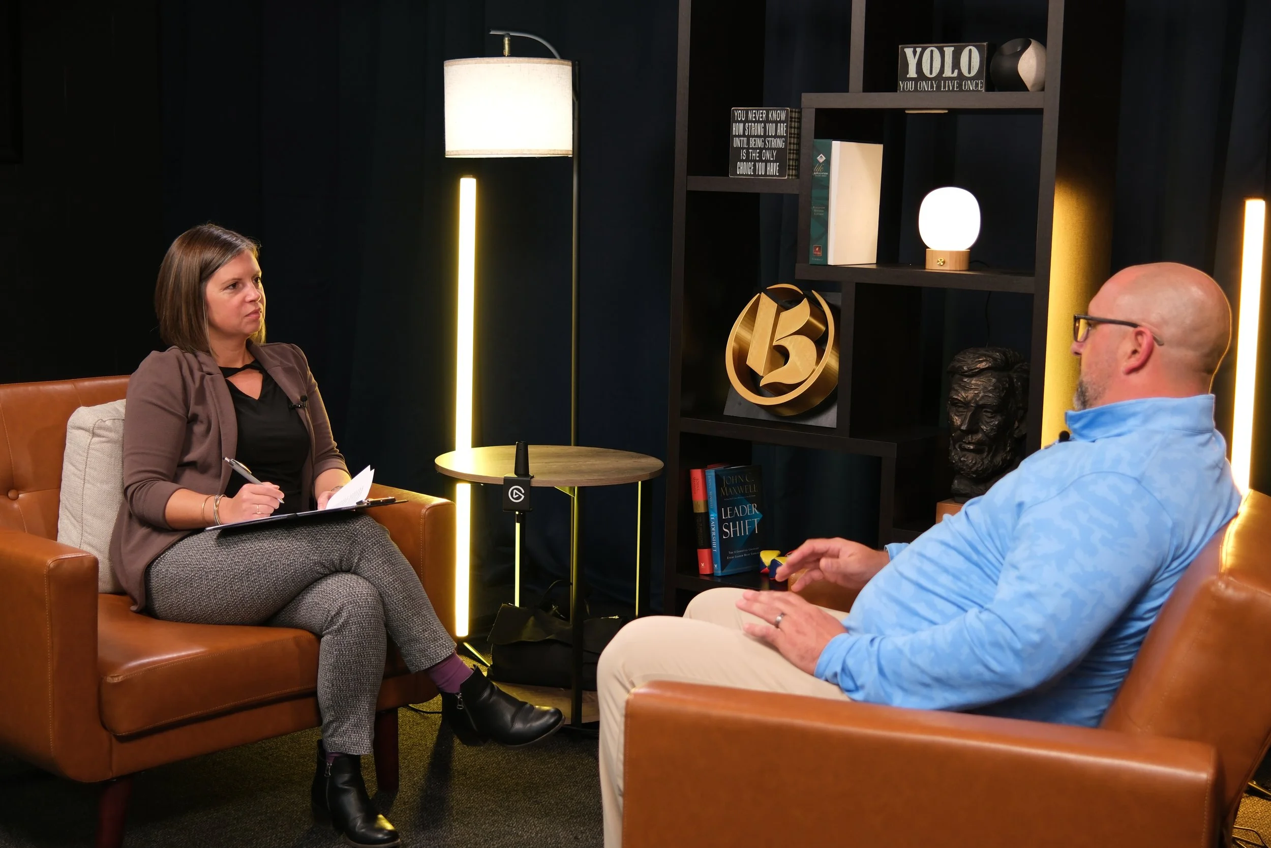 A woman interviewee sitting on a brown leather sofa, taking notes, and a man interviewer sitting on a similar sofa, in a studio with dark background, decorative shelves, books, and lighting.