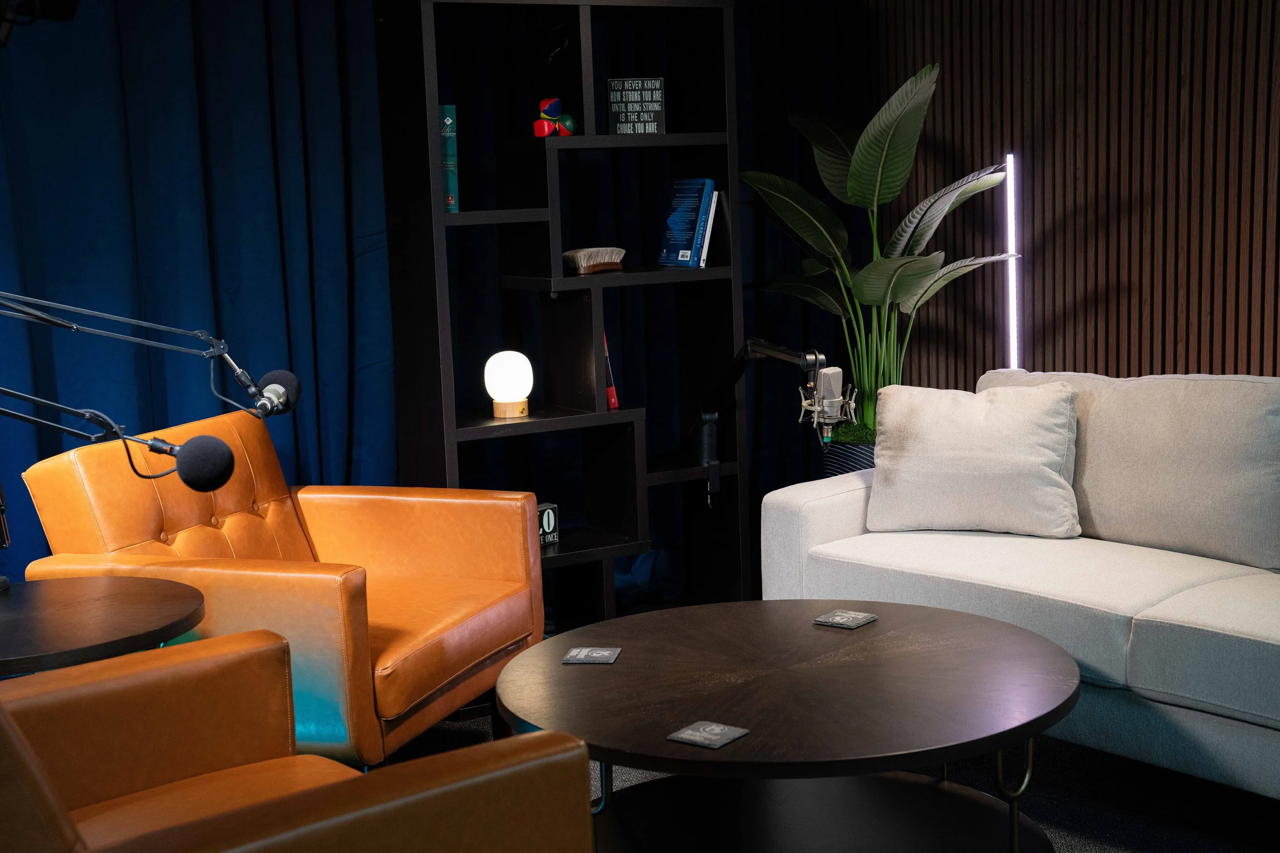 A cozy modern podcast recording room with a beige sofa, a brown leather armchair, a black coffee table with coasters, a black shelf with books and a small decorative lamp, a large plant with broad green leaves, a microphone, and a dark wood wall with a pink neon light vertical strip.